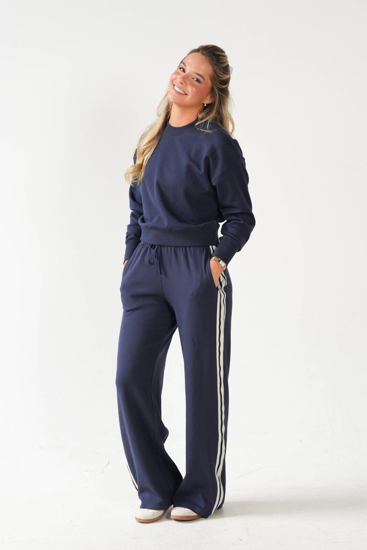 A woman modeling a matching navy blue sweatshirt and wide-leg sweatpants set with white side stripes, standing against a plain white studio background.