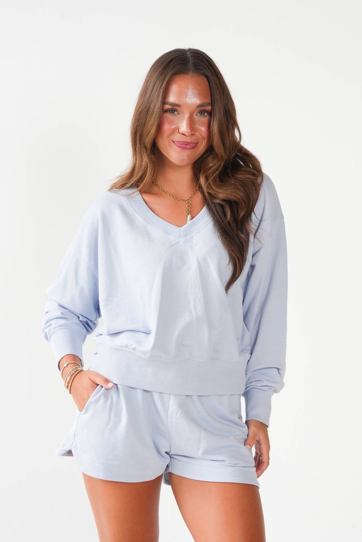 A woman modeling a light blue lounge set consisting of a long-sleeve V-neck sweatshirt and matching shorts, photographed against a clean white studio background.