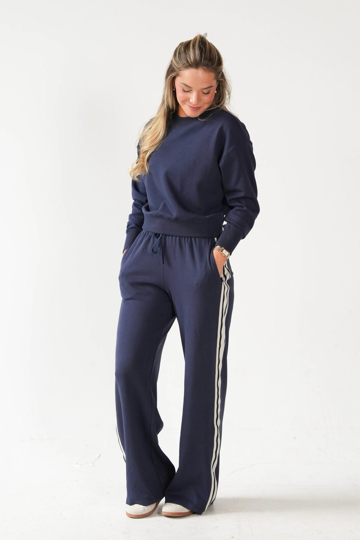 A woman modeling a matching navy blue sweatshirt and wide-leg sweatpants set with white side stripes, photographed against a clean white studio background.