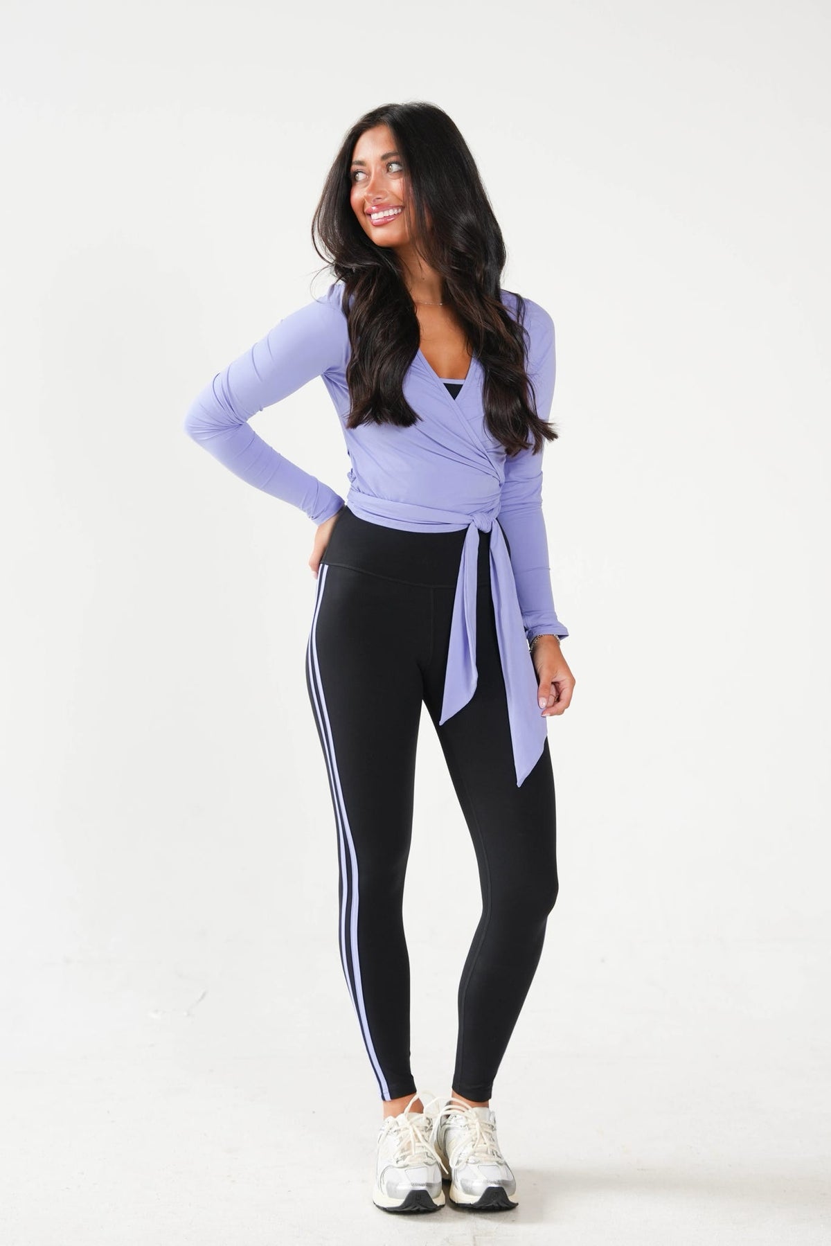 A woman modeling a light purple long-sleeve wrap top and black athletic leggings with white side stripes, paired with white sneakers, against a plain white studio background.