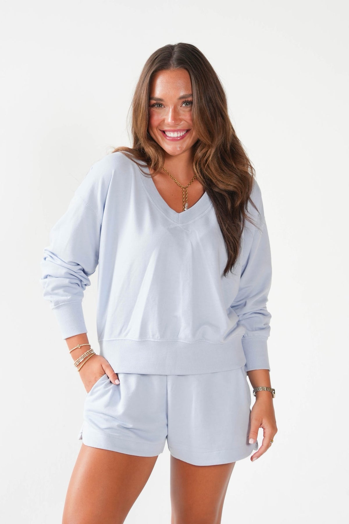 A woman modeling a light blue matching lounge set consisting of a long-sleeve V-neck sweatshirt and matching shorts, set against a plain white studio background.