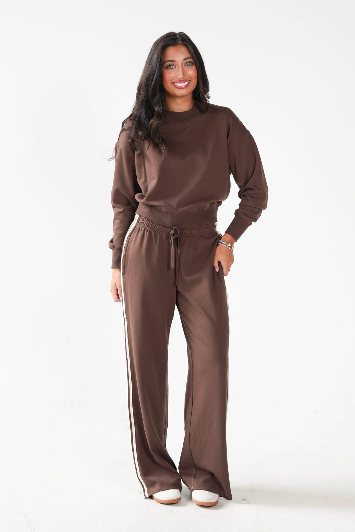 A woman modeling a matching brown lounge set consisting of a long-sleeve crewneck sweatshirt and wide-leg drawstring pants with white side stripes, photographed against a plain white studio background.
