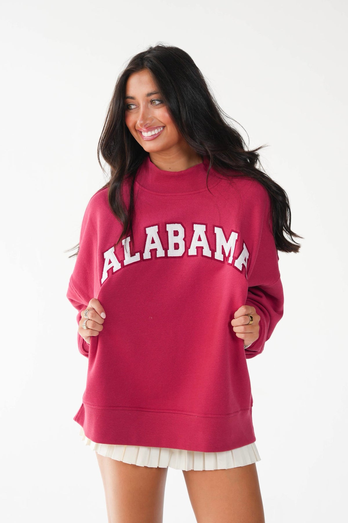 A woman wearing a bright pink crewneck sweatshirt with 'ALABAMA' printed in white arched letters across the chest, paired with a white pleated skirt, against a plain white background.