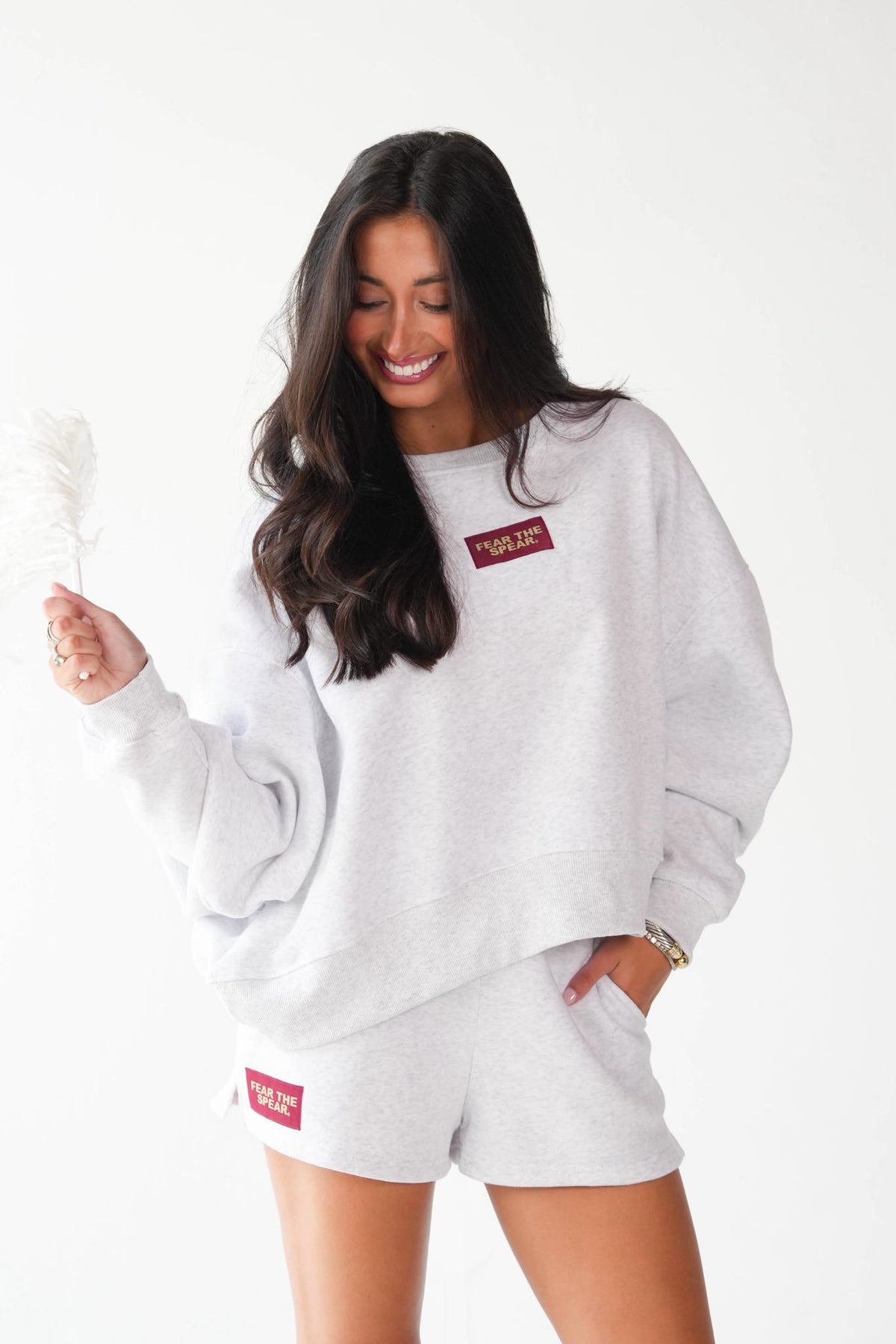 A woman modeling a light gray heather sweatshirt and matching shorts set, featuring a red rectangular patch with white text on the chest and thigh. She is holding a white feather against a clean white background.