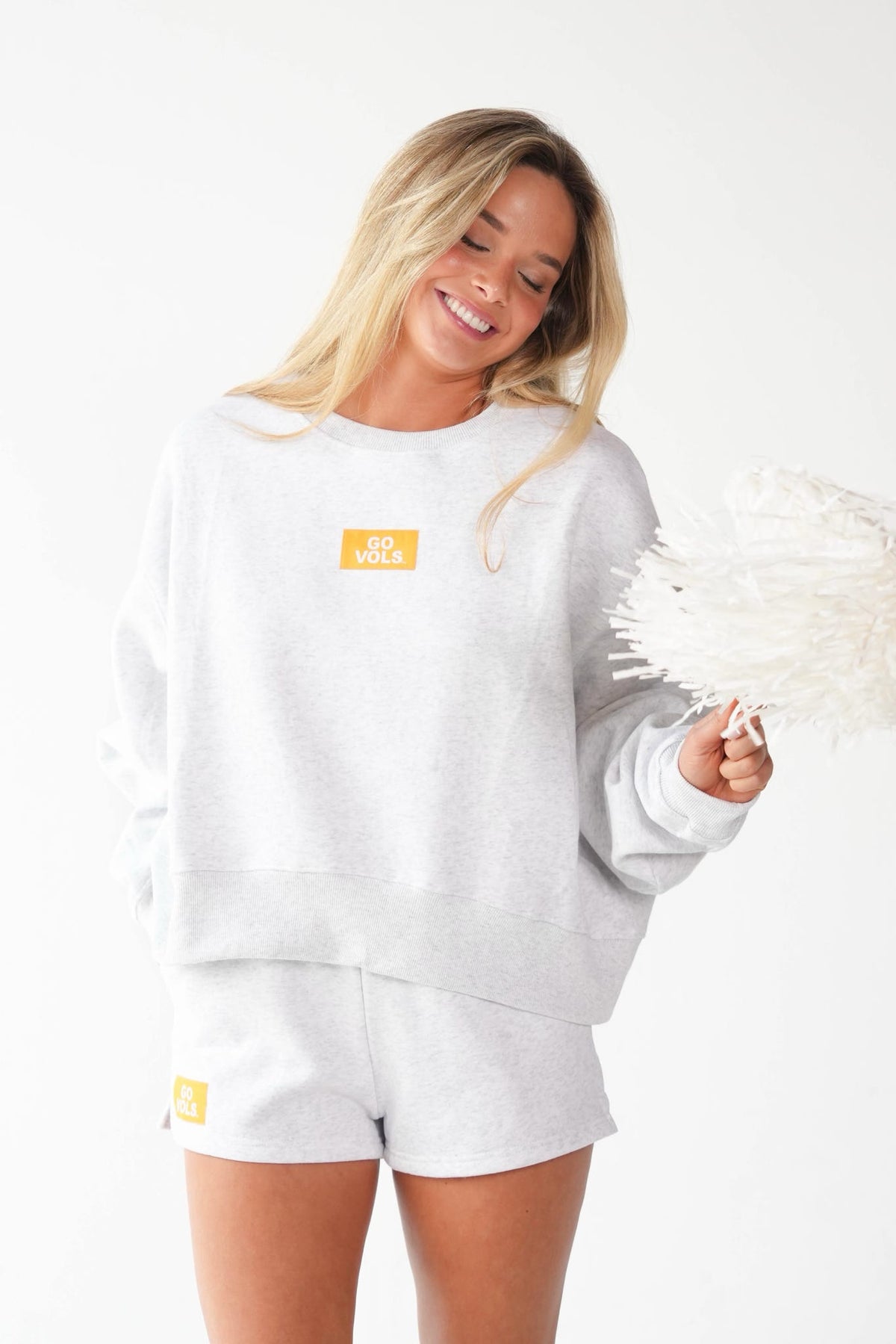 A woman wearing a light gray heather sweatshirt and matching shorts, holding a white feather duster against a plain white background. The sweatshirt and shorts feature a small yellow square logo on the chest and left leg.
