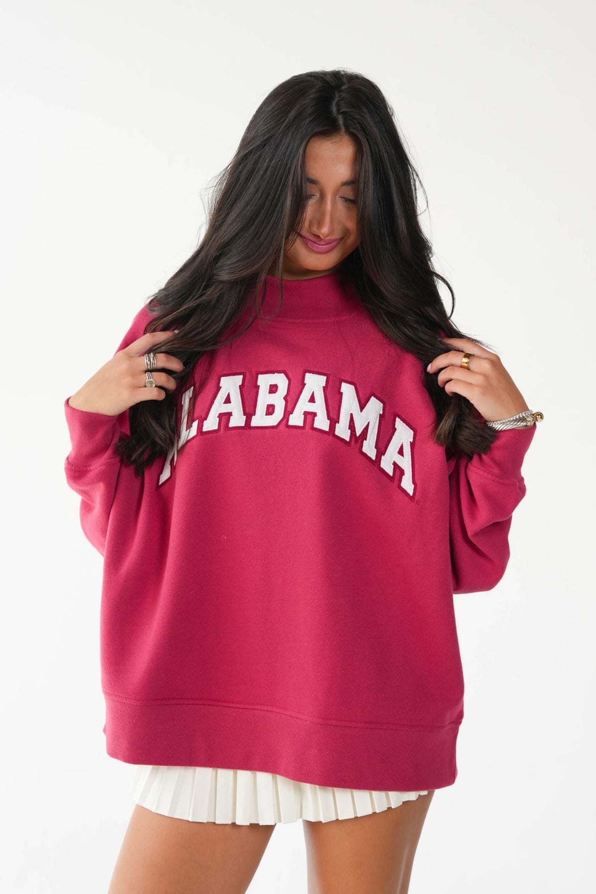 A woman wearing an oversized magenta sweatshirt with 'ALABAMA' printed in white arched letters across the chest, paired with a white pleated skirt, against a plain white background.