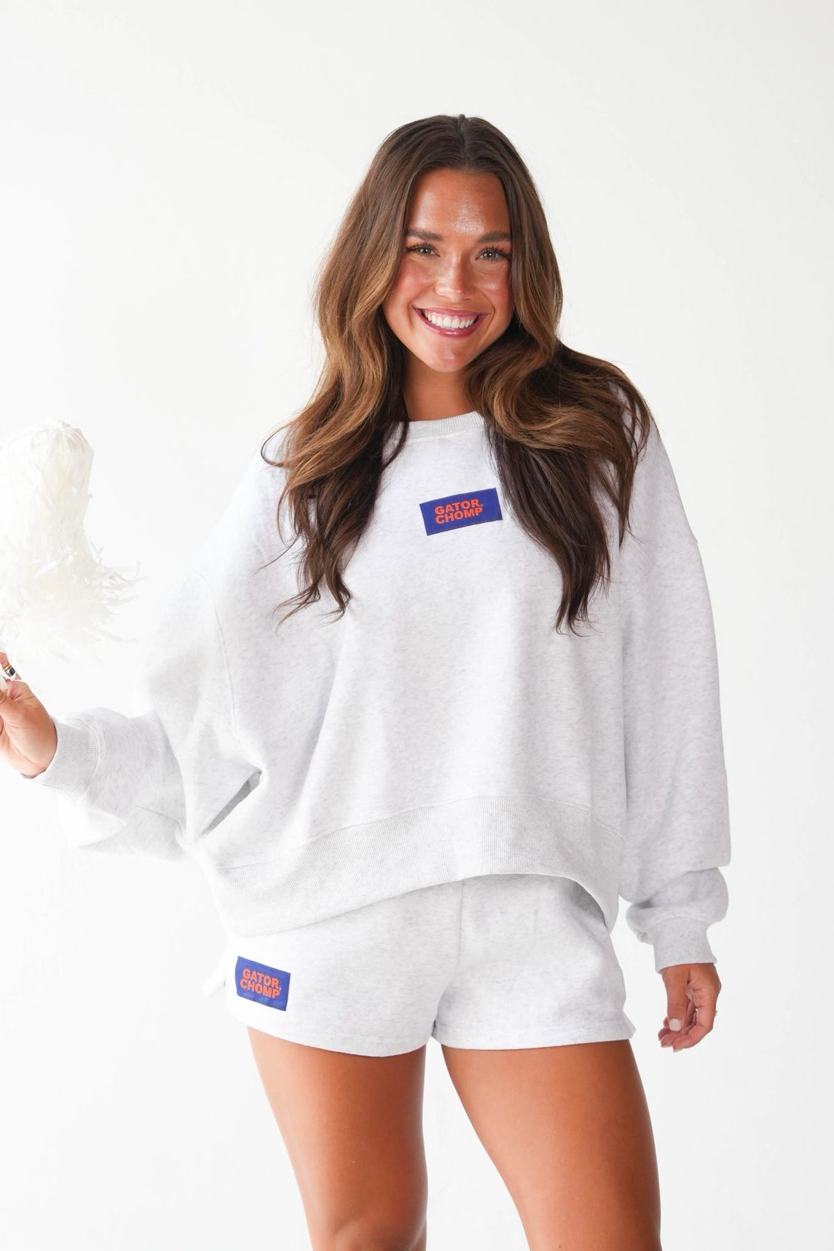 A woman modeling a white heathered sweatshirt and matching shorts set, featuring a small blue and red rectangular logo patch on the chest and shorts. She is holding a white feather wand against a clean white studio background.