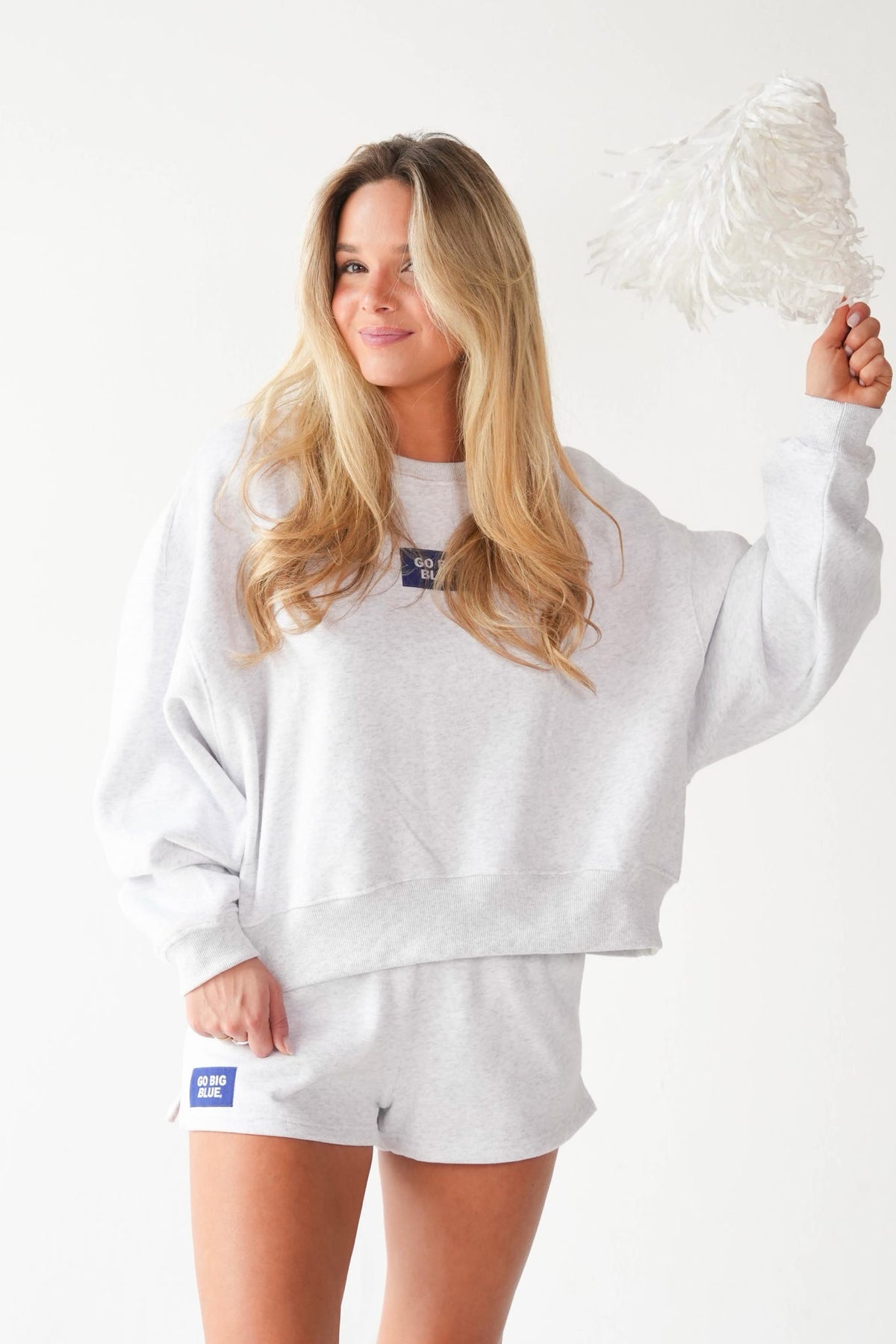 A woman modeling a light gray sweatshirt and matching shorts set, holding a white feather duster against a plain white background.