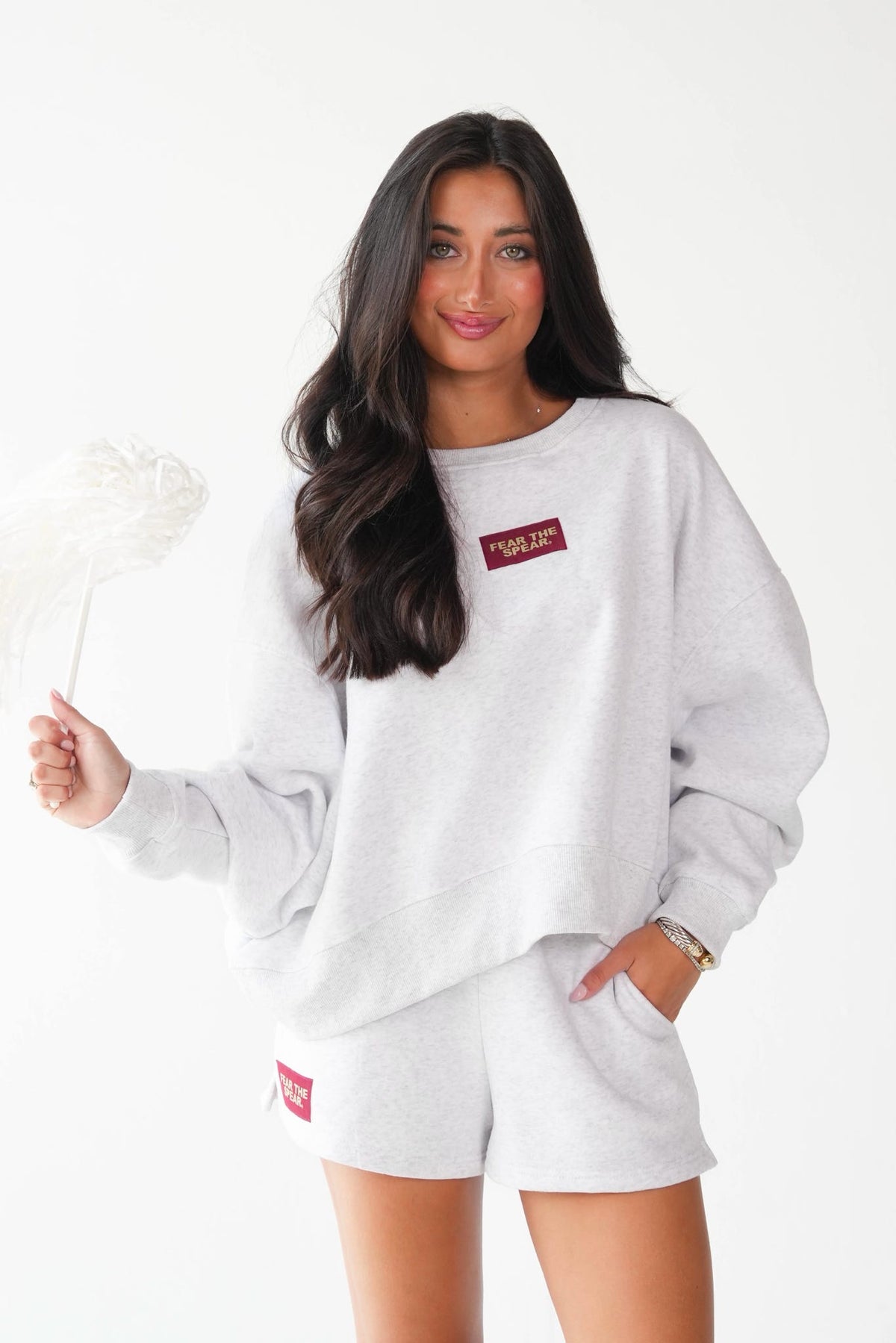 A woman modeling a light heather gray sweatshirt and matching shorts set, featuring a small red rectangular logo patch on the chest and shorts. She is holding a white feather fan against a clean white background.