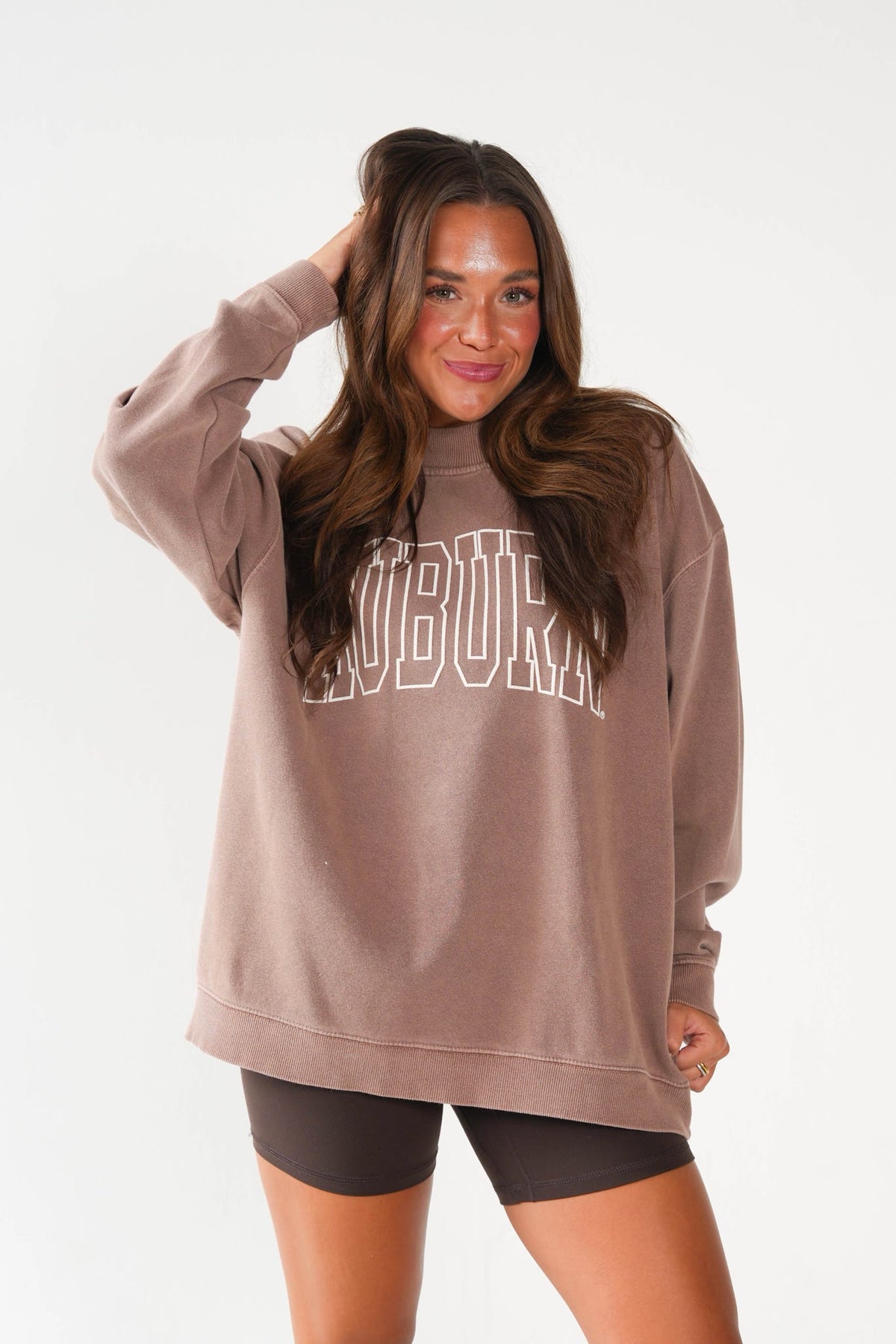 A woman modeling an oversized taupe sweatshirt with 'AUBURN' printed in white outline block letters across the chest, paired with black bike shorts against a plain white studio background.