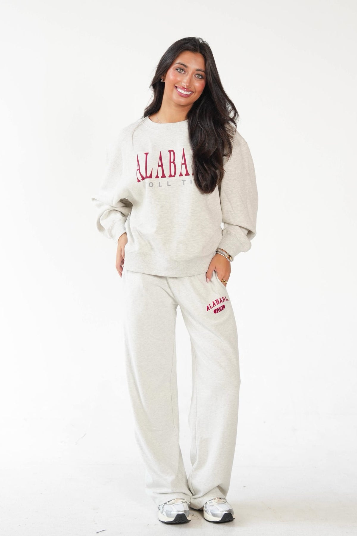 A woman modeling a matching sweatshirt and sweatpants set in a light heather gray color, featuring 'ALABAMA' printed in red on the chest and thigh.