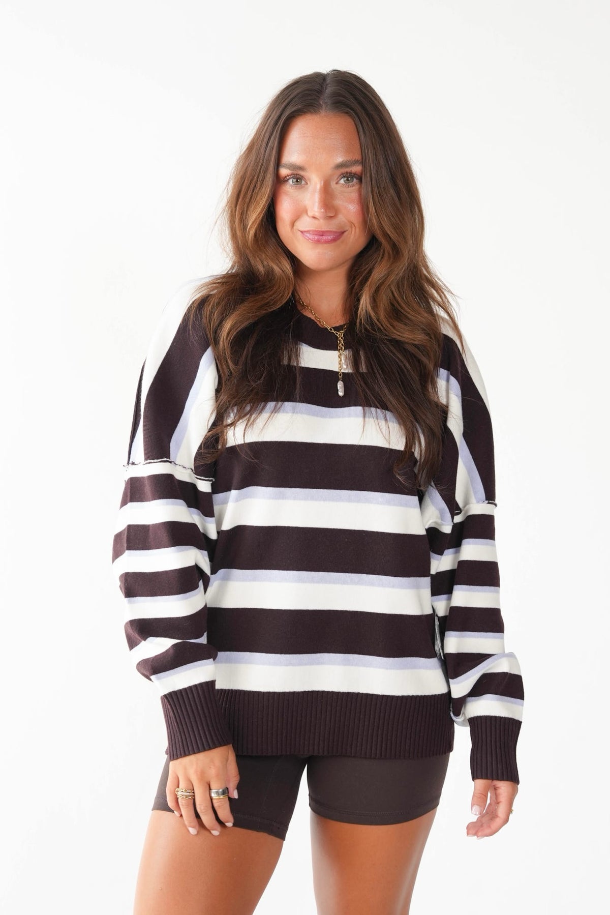 A woman modeling a dark brown and white horizontally striped knit sweater with a mock neck and ribbed cuffs, paired with dark brown shorts against a plain white background.