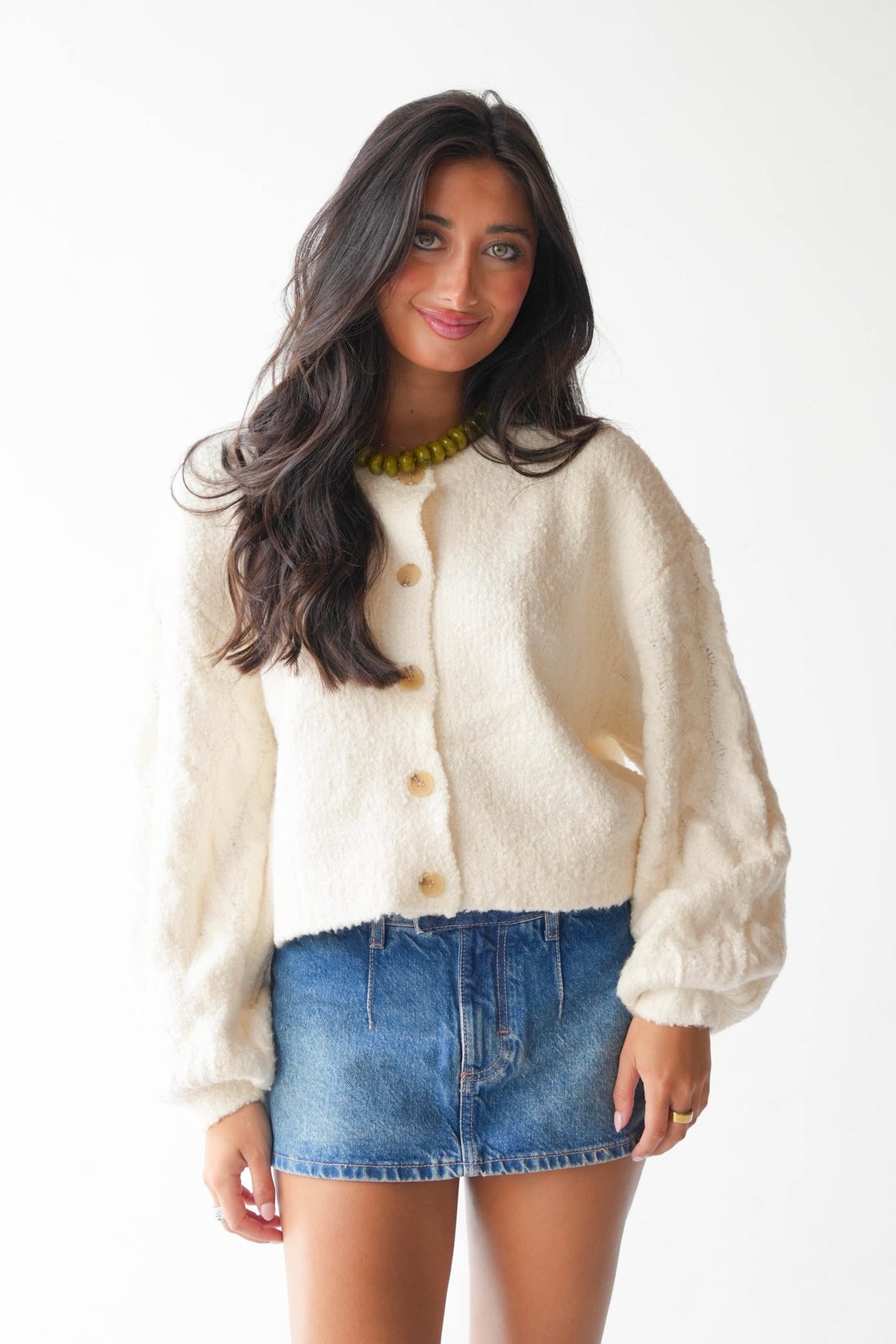 A woman modeling a cream-colored, fuzzy cropped cardigan with gold buttons, paired with a blue denim mini skirt and a green beaded necklace, against a plain white background.