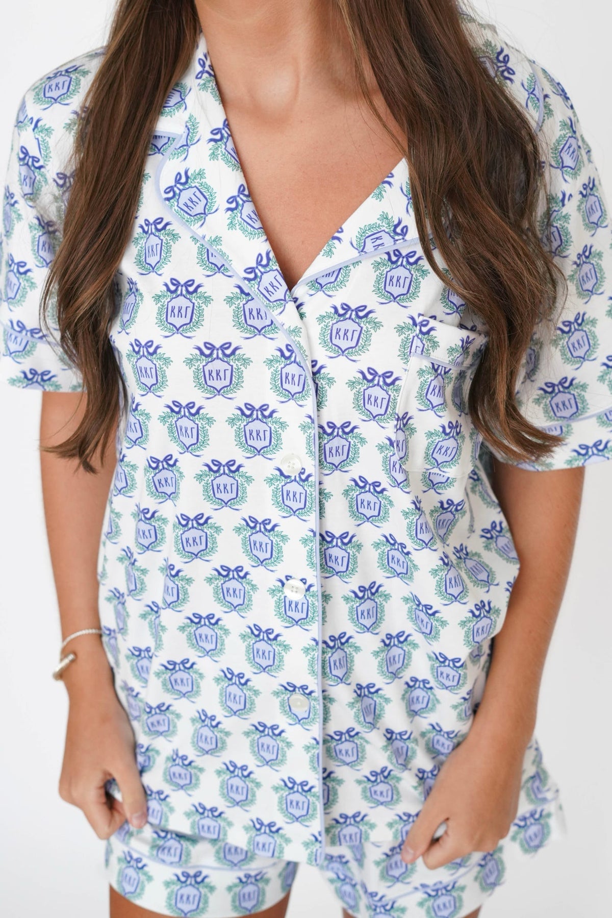 A woman wearing a white short-sleeve button-front romper or pajama-style top with an allover blue and green monogram print. The image is cropped from the neck to the upper thighs, showing the garment's fit and pattern against a plain light background.