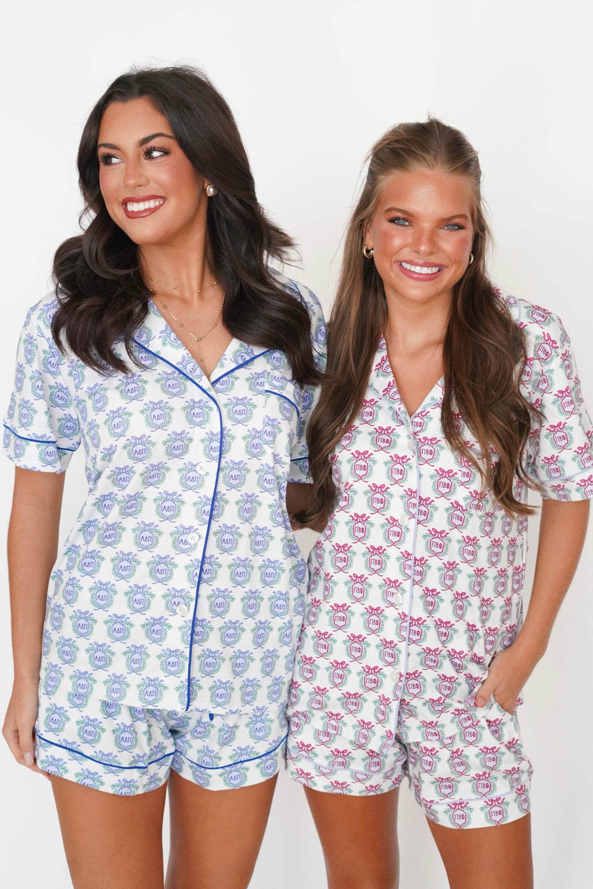 Two women modeling matching short-sleeve pajama sets against a plain white background. The set on the left is white with a blue floral pattern and blue piping, and the set on the right is white with a pink floral pattern and pink piping.