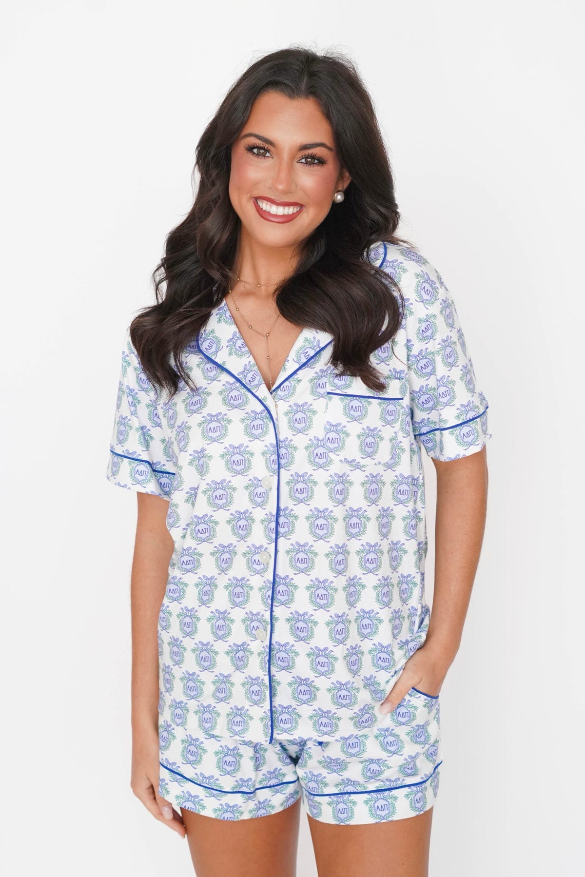 A woman modeling a matching short-sleeve pajama set with a blue and white repeating crest pattern and blue piping, photographed against a plain light background.