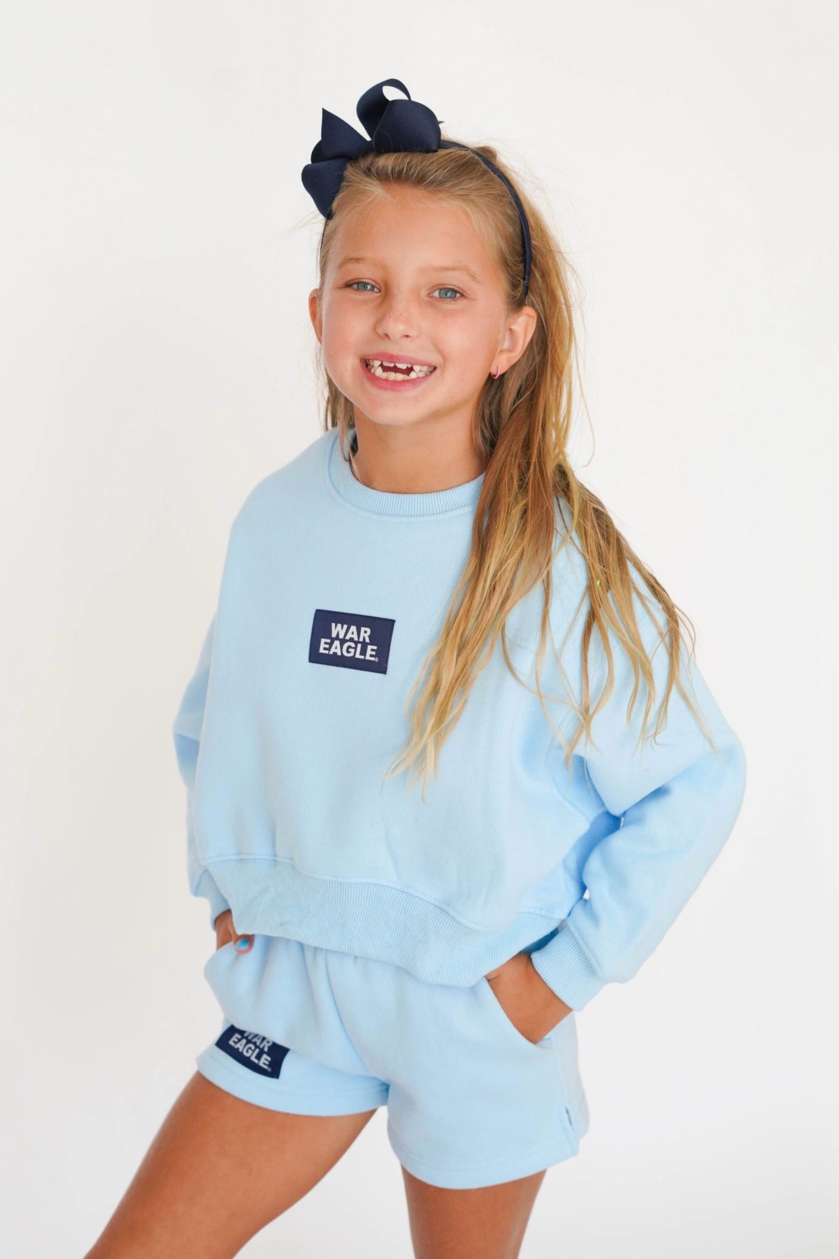 A young girl with a black hair bow is wearing a light blue sweatshirt and matching shorts set with a navy blue 'WAR EAGLE' patch on the chest, posed against a plain white background.