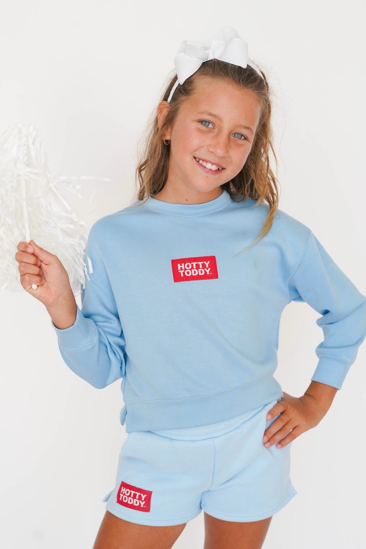 A young girl smiling and wearing a light blue long-sleeve sweatshirt and matching shorts, both featuring a red square patch with the text 'HOTTY TODAY' in white. She is posing against a plain white background, holding a crumpled plastic bag.