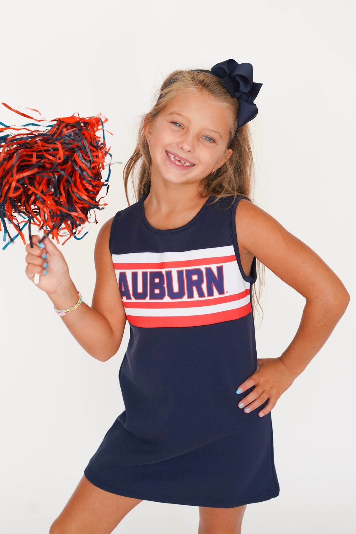 A young girl smiling and posing in a navy blue sleeveless cheerleader dress with 'AUBURN' printed across the chest in white and red stripes. She is holding a red and navy pom-pom and wearing a navy hair bow against a plain white background.