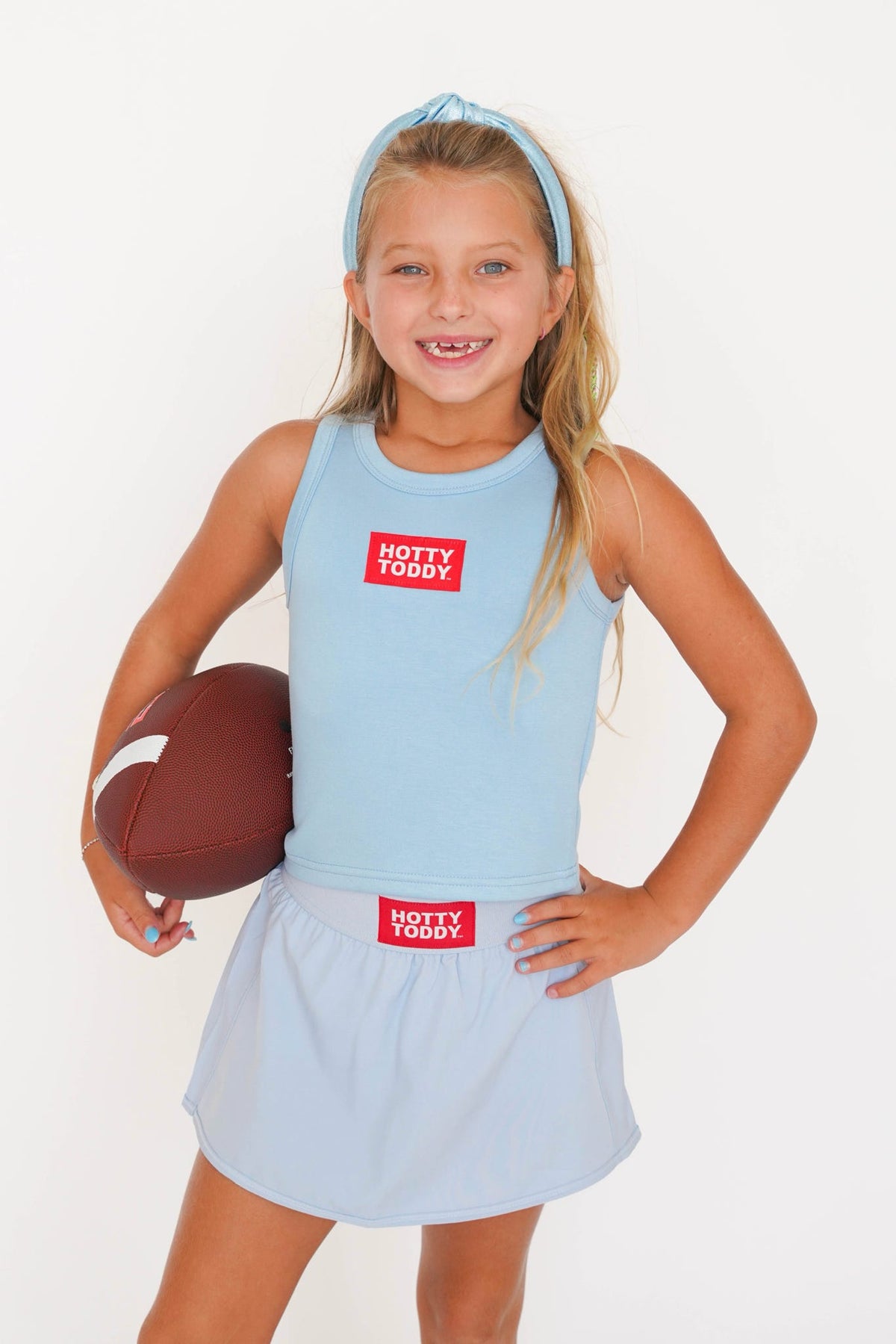 A young girl with blonde hair wearing a light blue athletic tank top and matching skirt, holding a brown football against a plain white background. The top and skirt feature a red square patch with the text 'HOTTY TODDY'.