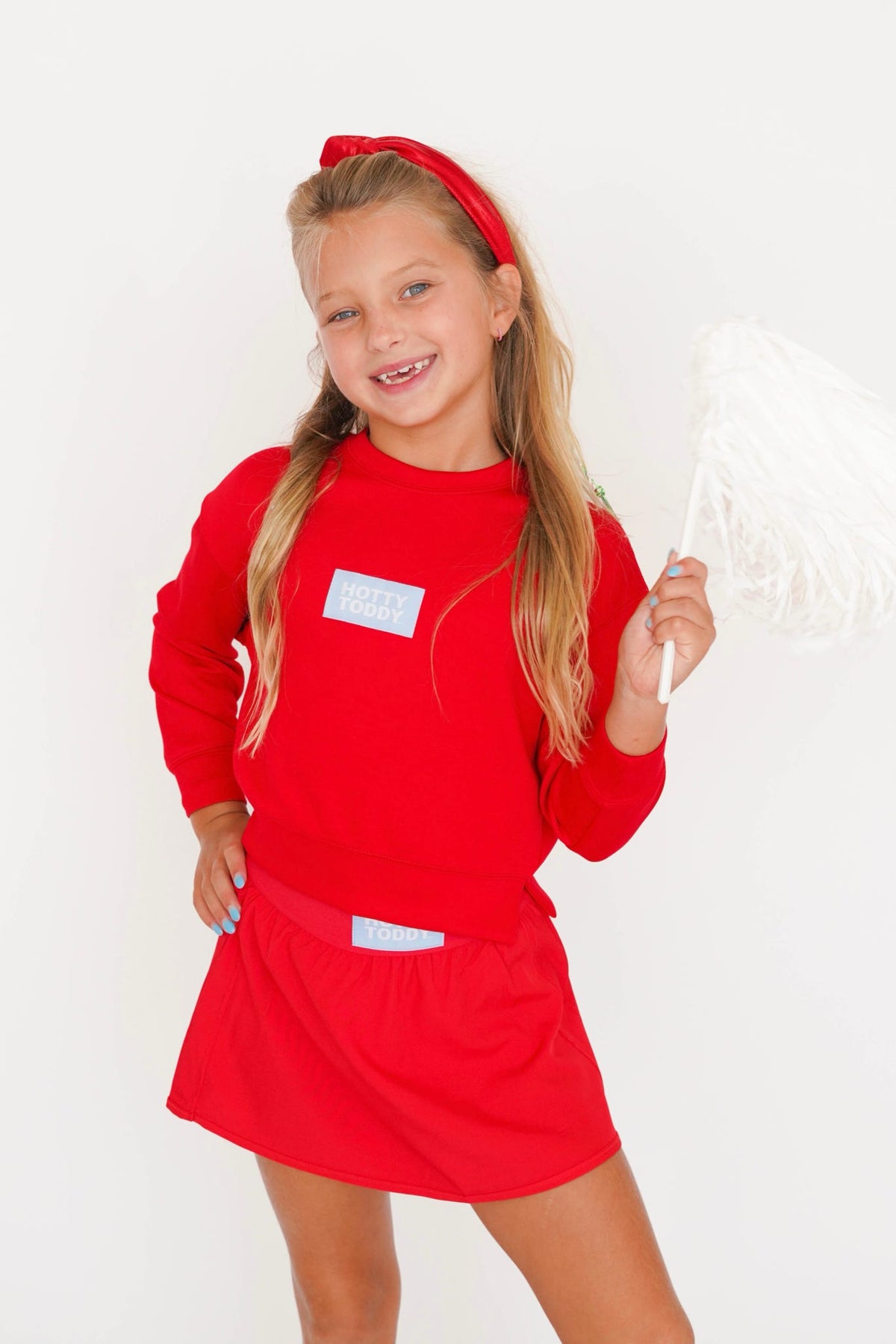 A young girl smiling and posing in a bright red cheerleader-style outfit consisting of a long-sleeve top, matching skirt, and red headband, while holding a white pom-pom against a plain white background.