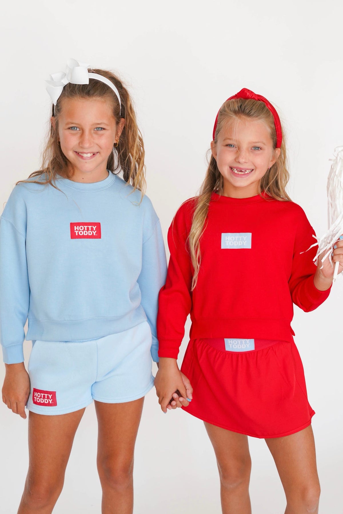 Two young girls modeling matching athletic sets. The girl on the left wears a light blue sweatshirt and shorts with a red chest patch reading 'HOBBY TEAM'.