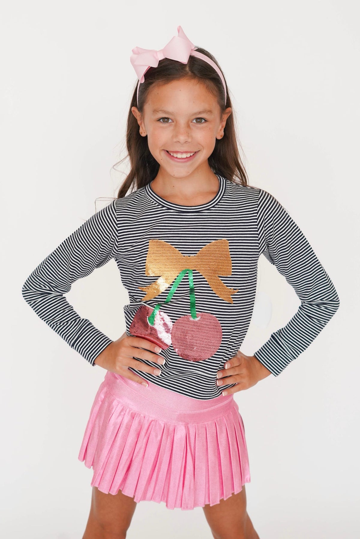 A young girl smiling and posing in a black and white striped long-sleeve top with a sequined cherry and bow graphic, paired with a pink pleated skirt and a pink bow headband, against a plain white background.