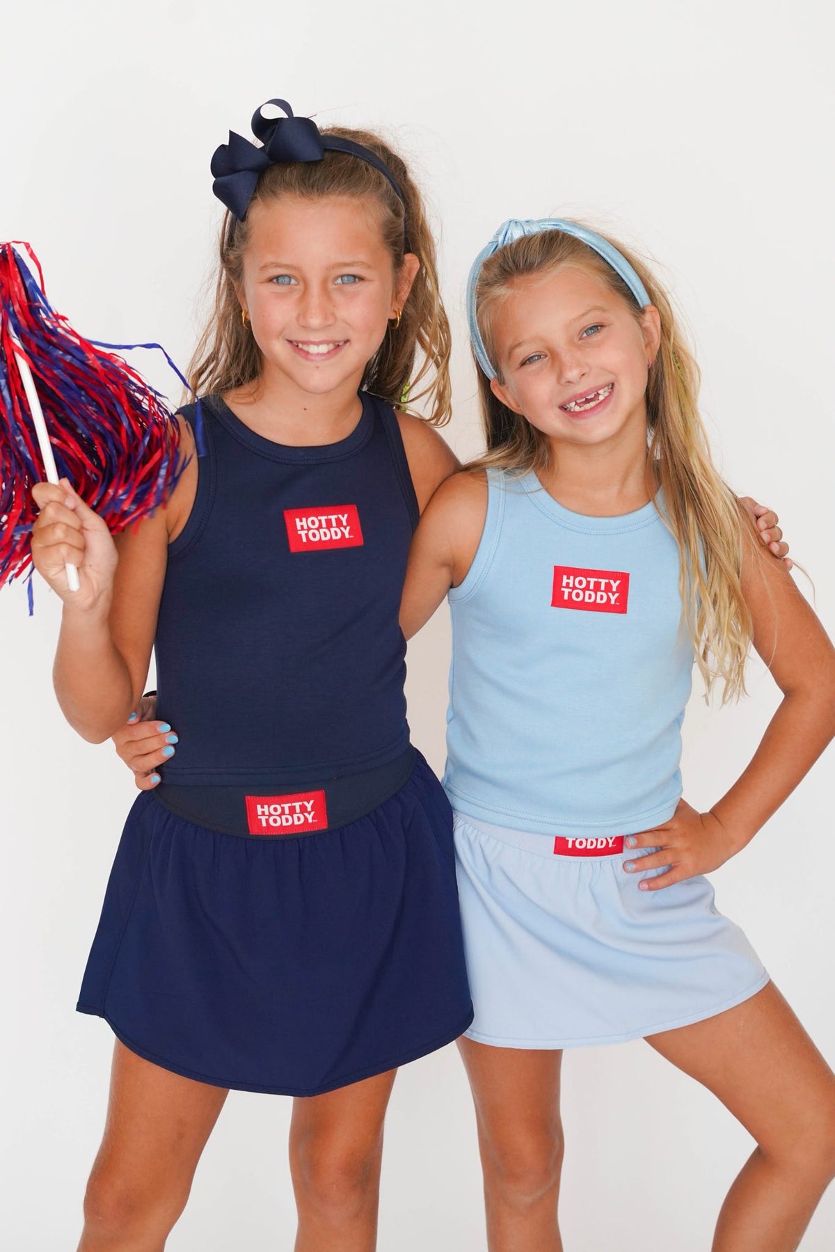 Two young girls modeling matching athletic tank tops and shorts sets. The girl on the left wears a navy blue set and holds a red and blue pom-pom, while the girl on the right wears a light blue set.