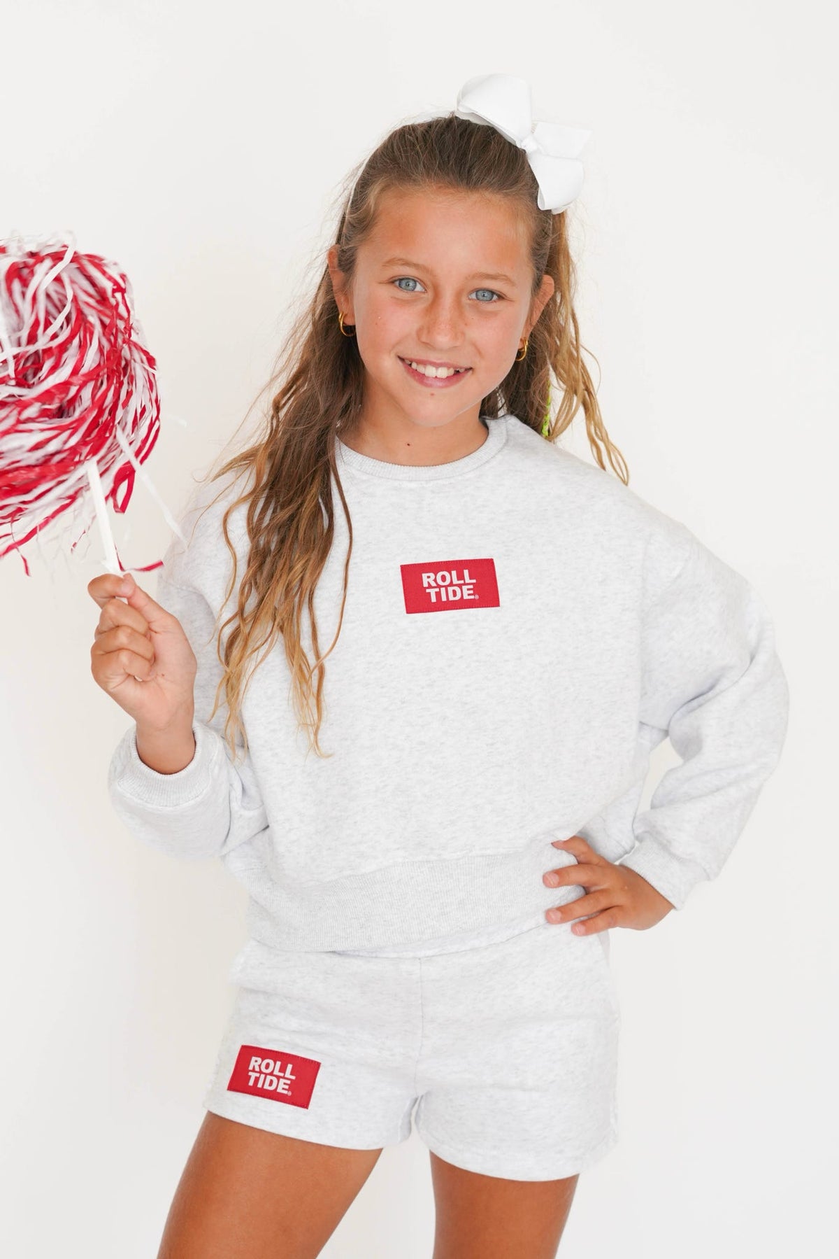 A young girl smiling and holding a red and white striped pom-pom, wearing a light heather gray sweatshirt and matching shorts with red 'ROLL TIDE' logo patches on the chest and shorts.