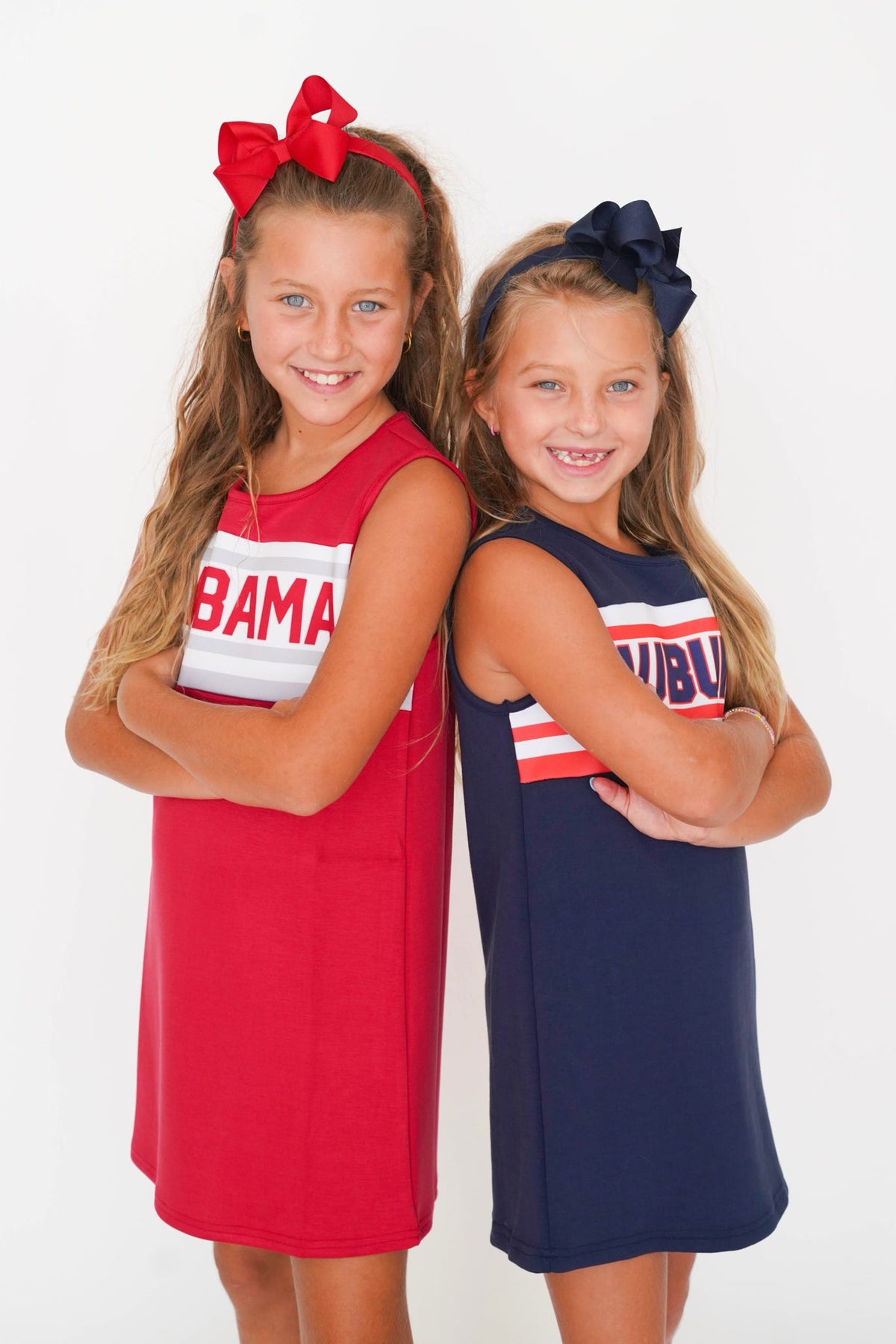 Two young girls modeling sleeveless athletic dresses with 'Alabama' text, standing side-by-side against a white studio background. The girl on the left wears a red dress with a white stripe and a red bow headband.