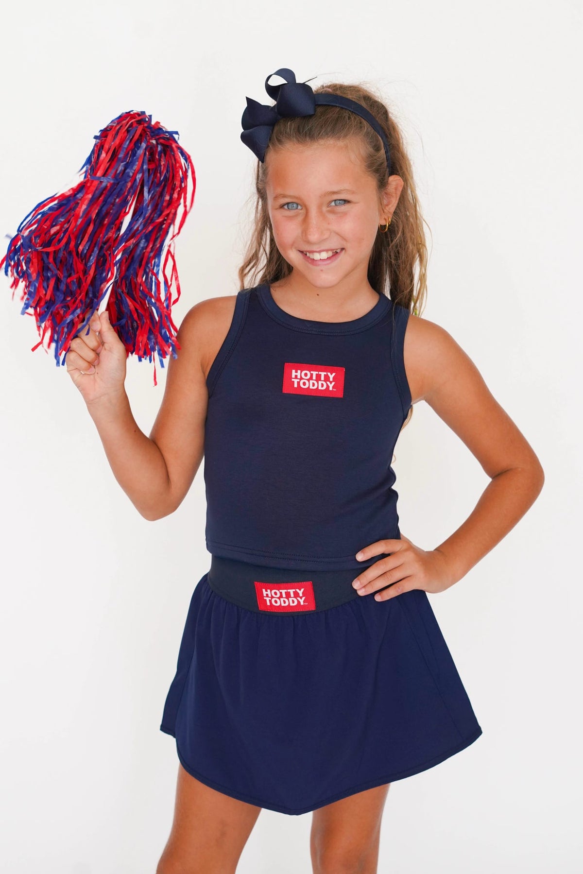 A young girl cheerleader models a navy blue sleeveless tank top and matching skirt set with red and white 'HOTTY TODDY' brand patches. She holds a red and blue pom-pom against a clean white background.