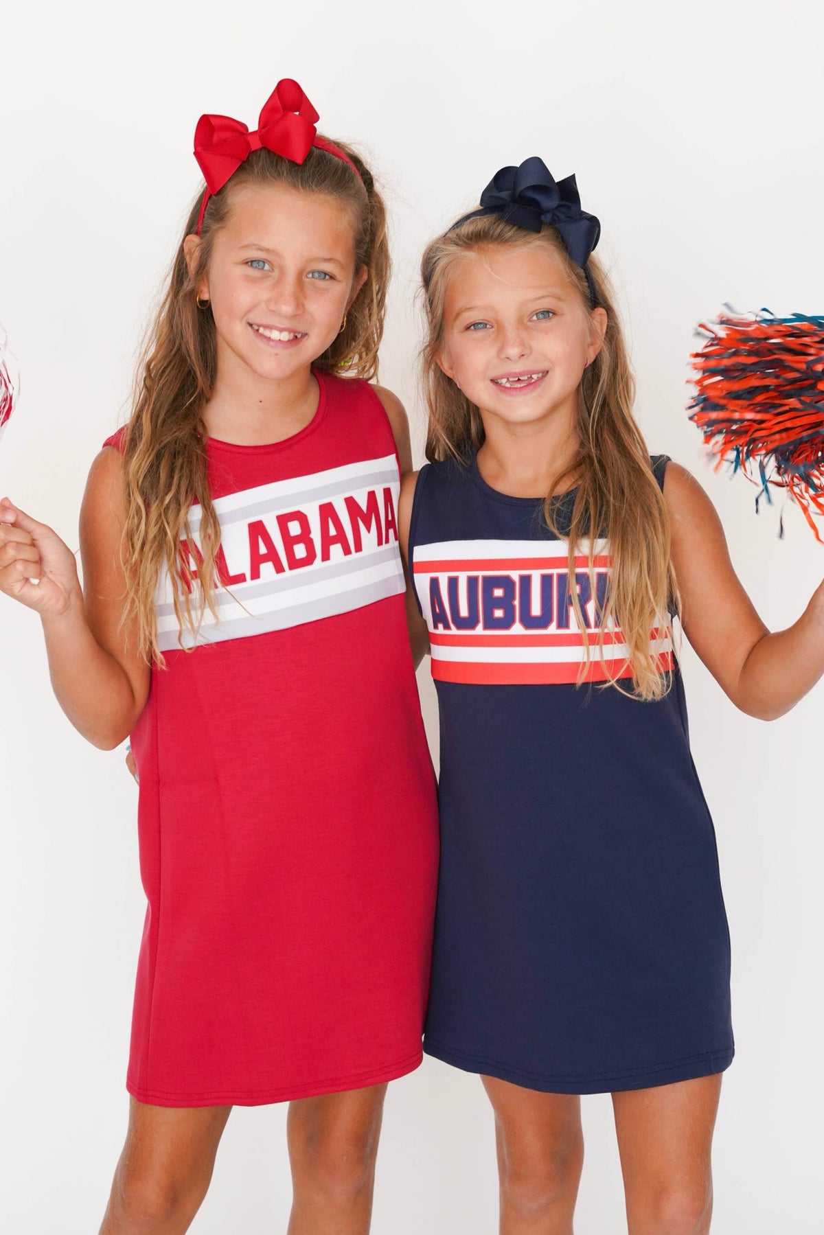 Two young girls modeling sleeveless Alabama Auburn cheerleader dresses. The girl on the left wears a red dress with a white stripe and 'ALABAMA' text, paired with a red bow.