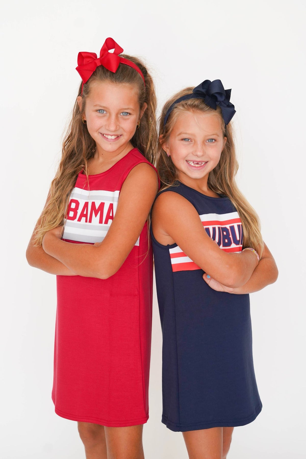 Two young girls modeling sleeveless Alabama-themed dresses, standing side-by-side against a plain white studio background. The girl on the left wears a red dress with a white stripe and 'ABAMA' text, paired with a red bow headband.