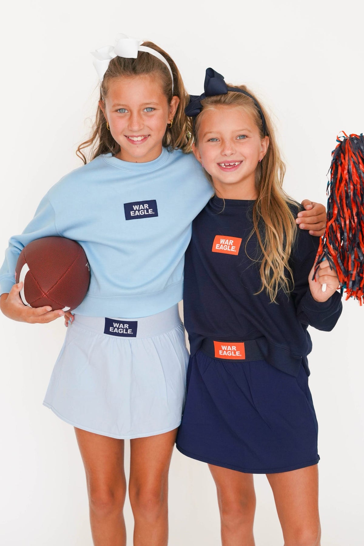 Two young girls modeling matching navy and light blue long-sleeve t-shirts with 'WAR EAGLE' patches, paired with matching skirts, while one holds a football and the other holds pom-poms against a white studio background.