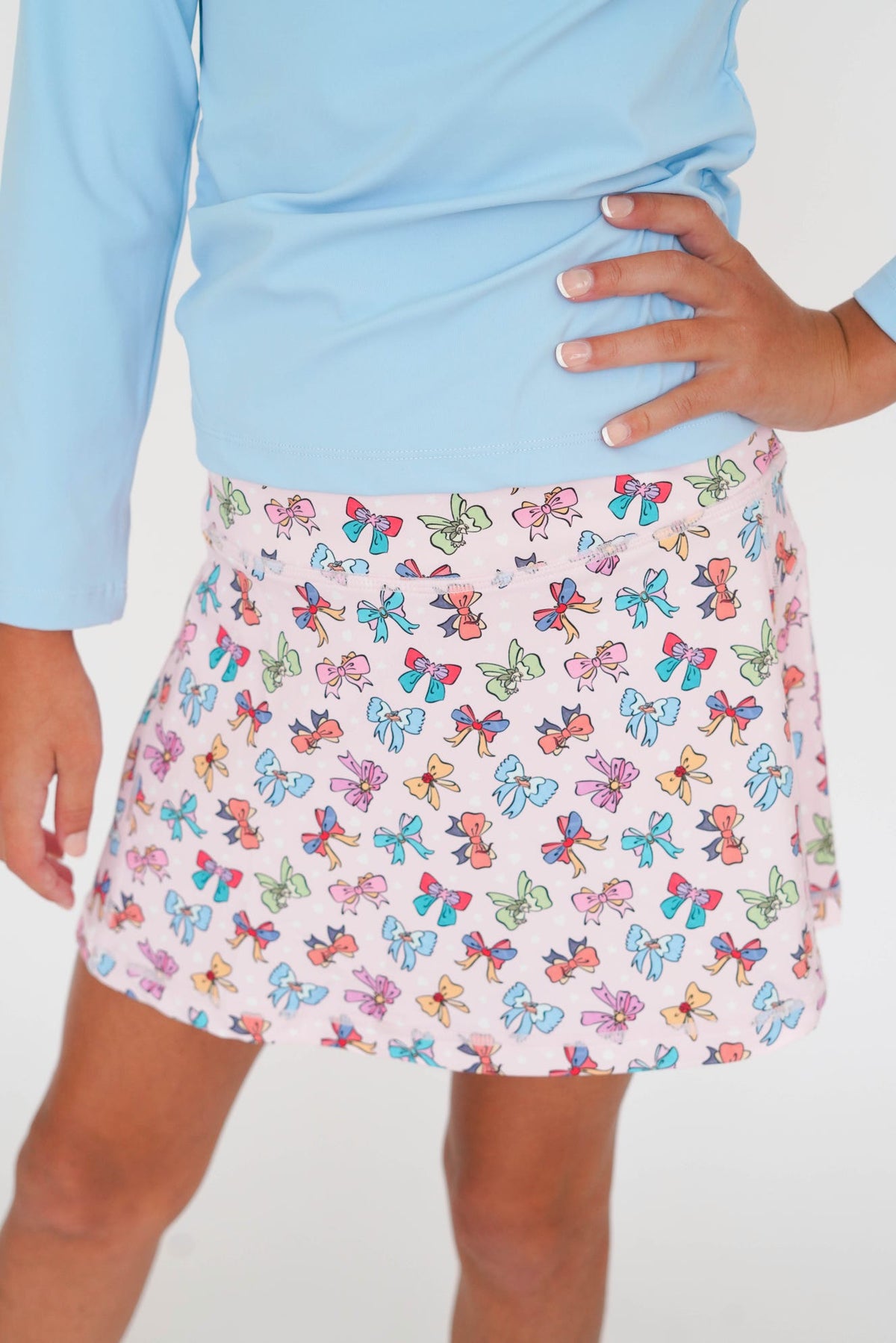 A child wearing a light blue long-sleeve top and a pink skirt with a colorful butterfly print, shown from the waist down against a plain white background.
