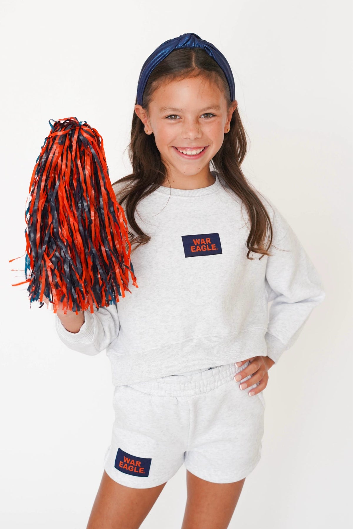 A young girl smiling and holding a red and navy blue pom-pom, wearing a light gray sweatshirt and matching shorts with 'WEAR EAGLE' patches on the chest and leg, against a plain white background.