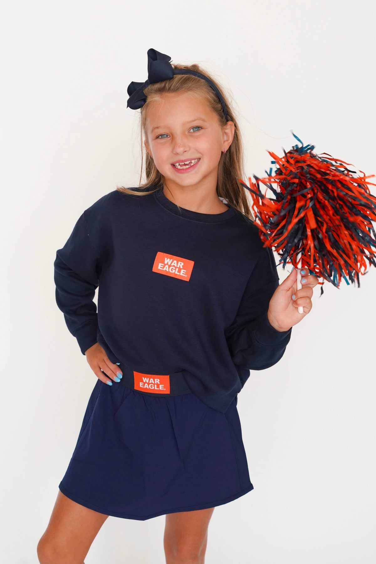 A young girl smiling and posing in a navy blue cheerleader outfit, featuring a long-sleeve sweatshirt with a red 'WAS EAGLE' patch and a matching skirt, while holding a red and navy pom-pom against a plain white background.