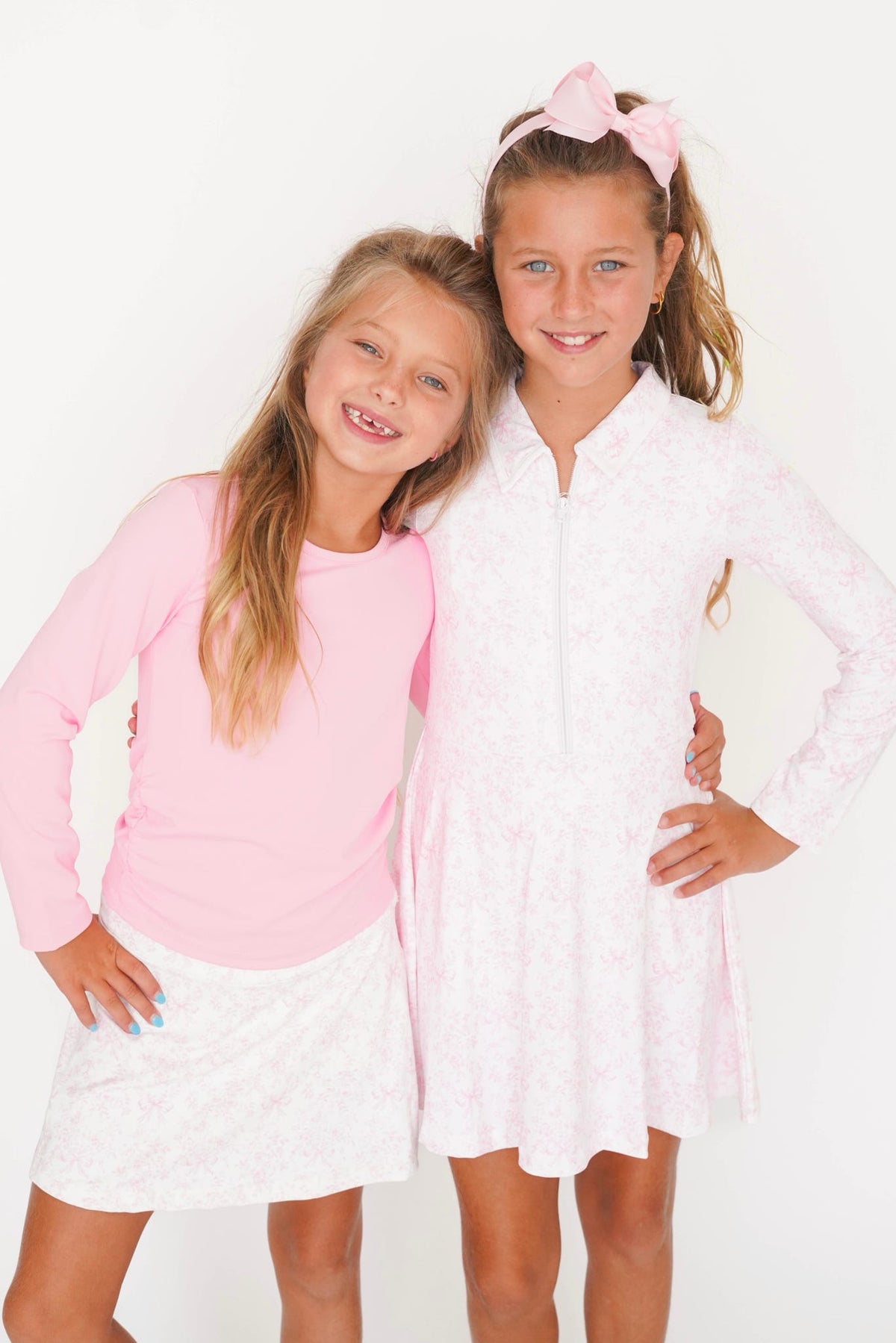 Two young girls posing together against a plain white background. The girl on the left wears a light pink long-sleeve top and a white patterned skirt, while the girl on the right wears a white patterned collared dress with a pink bow in her hair.