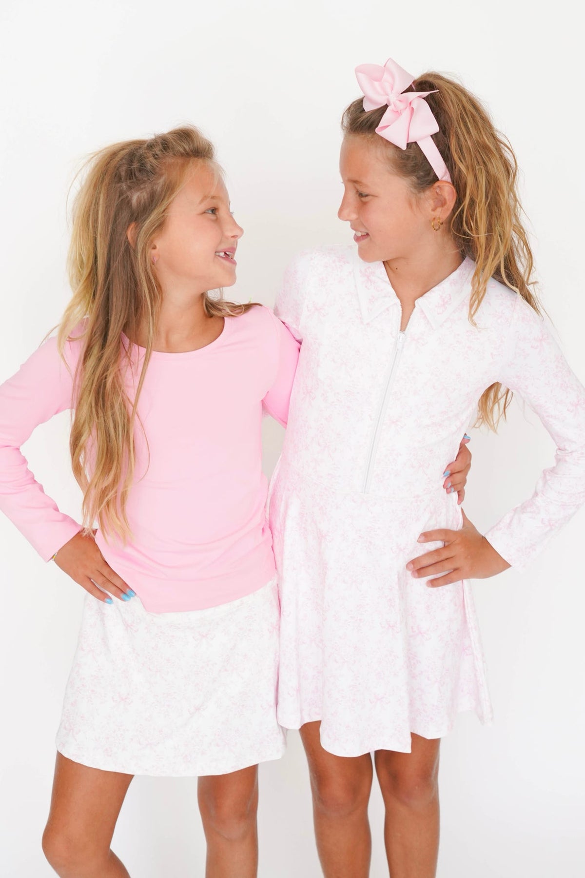 Two young girls standing and smiling at each other against a plain white studio background. The girl on the left wears a pink long-sleeve top and a white skirt, while the girl on the right wears a white long-sleeve dress with a pink bow headband.