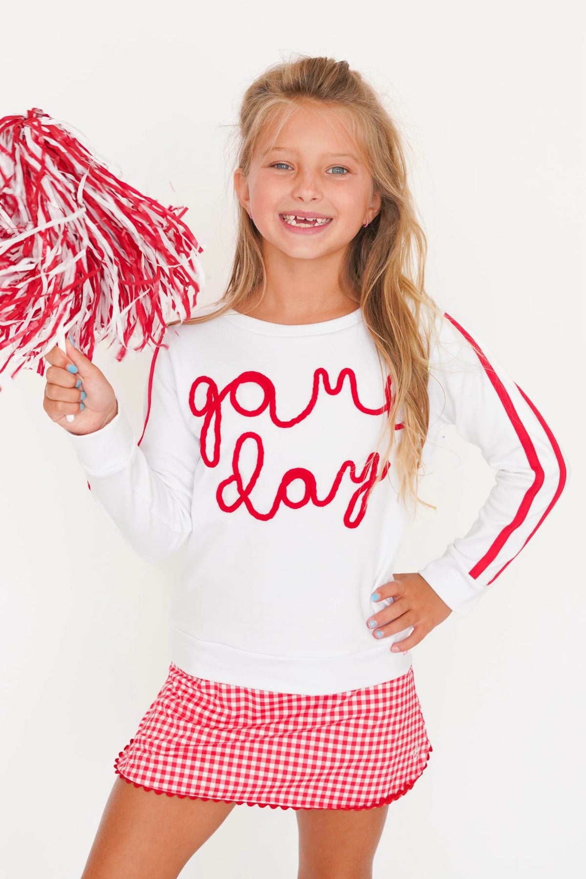 A young girl cheerleader posing in a white long-sleeve sweatshirt with red script text reading 'game day' and red stripes on the sleeves, paired with a red and white gingham mini skirt and holding a red and white pom-pom.