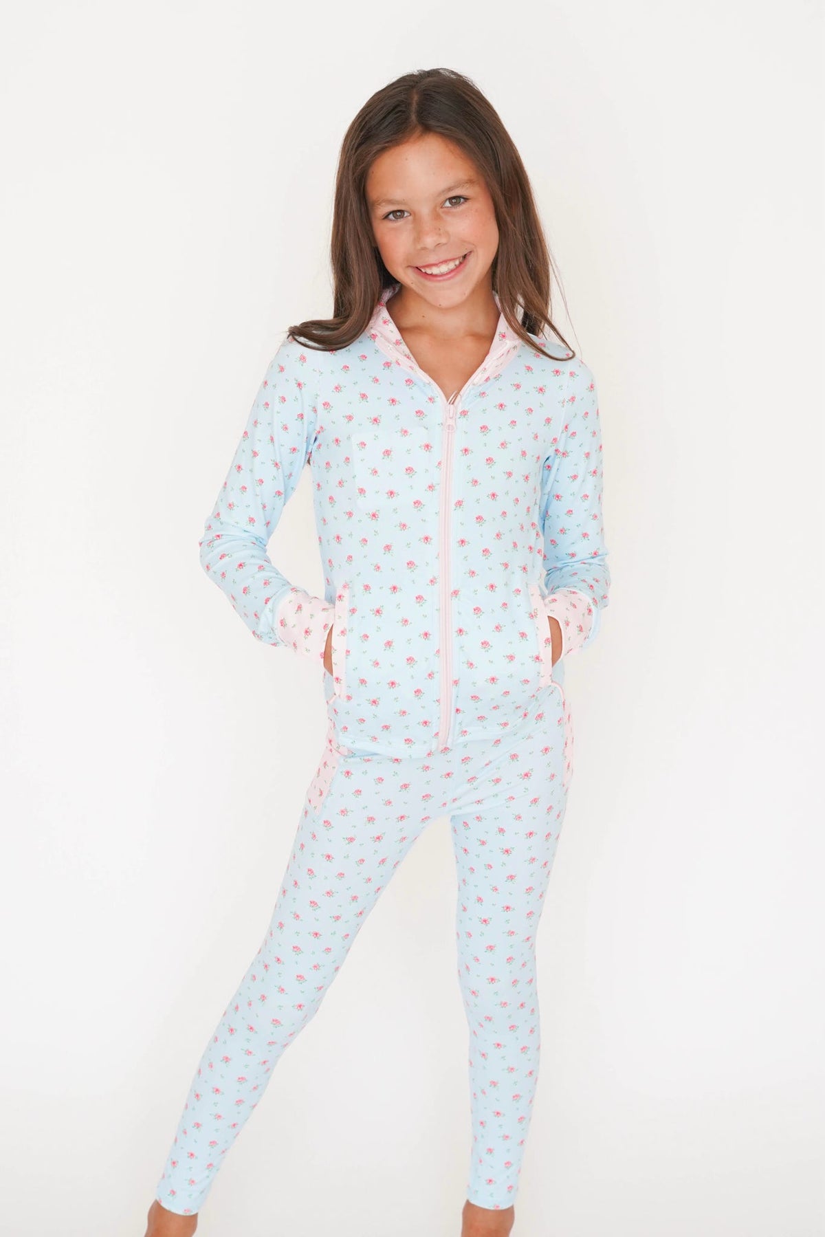 A young girl smiling while wearing a light blue long-sleeve zip-up pajama set with a pink and white pattern, photographed against a plain white background.