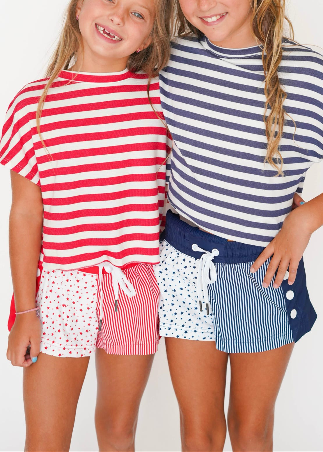 Two young girls smiling and posing together in matching nautical-themed striped t-shirts and shorts. The girl on the left wears a red and white striped shirt with pink polka-dot shorts, and the girl on the right wears a navy and white striped shirt with navy and white striped shorts.