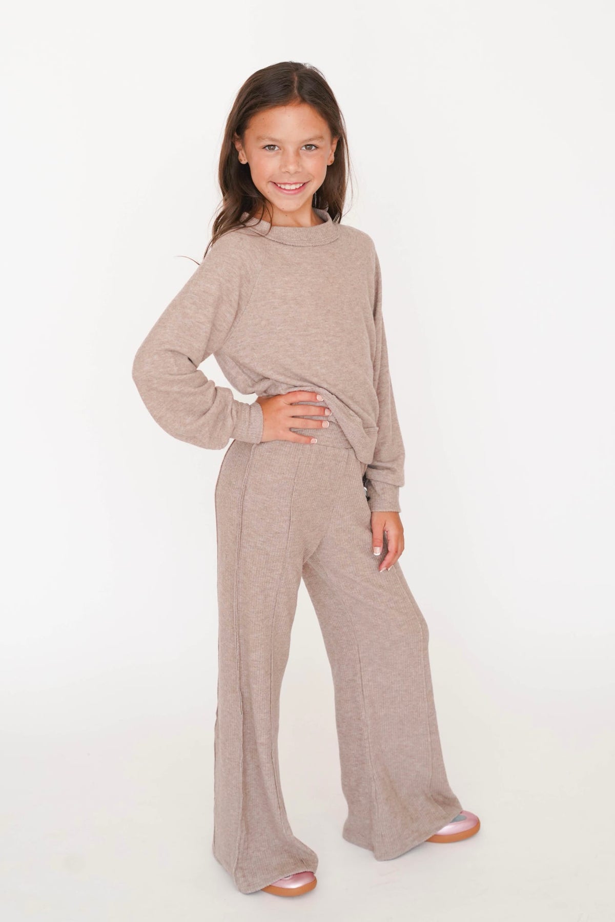 A young girl modeling a matching two-piece lounge set consisting of a long-sleeve mock neck top and wide-leg pants in a heathered taupe color, set against a plain white studio background.