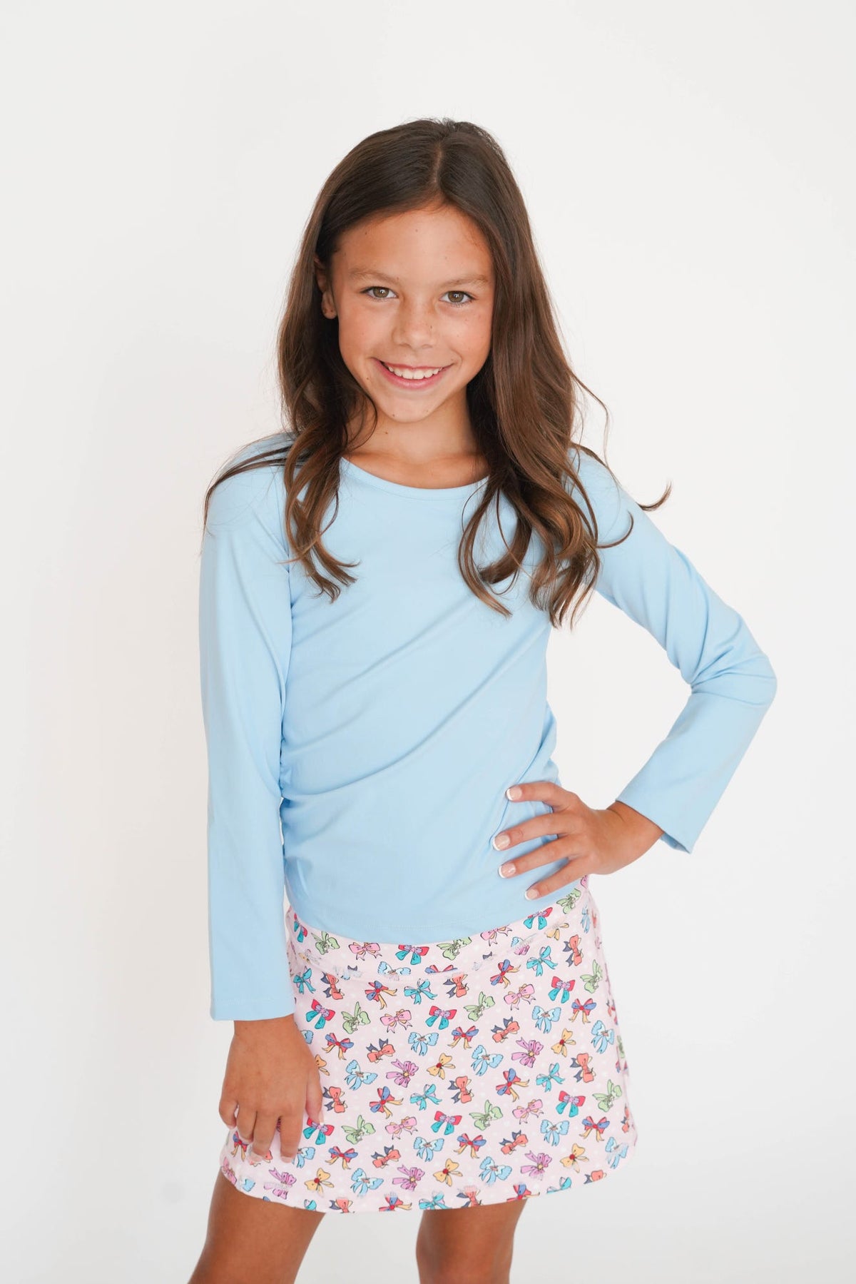 A young girl with long brown hair stands against a plain white background, wearing a light blue long-sleeve top and a white skirt with a colorful floral and butterfly print.