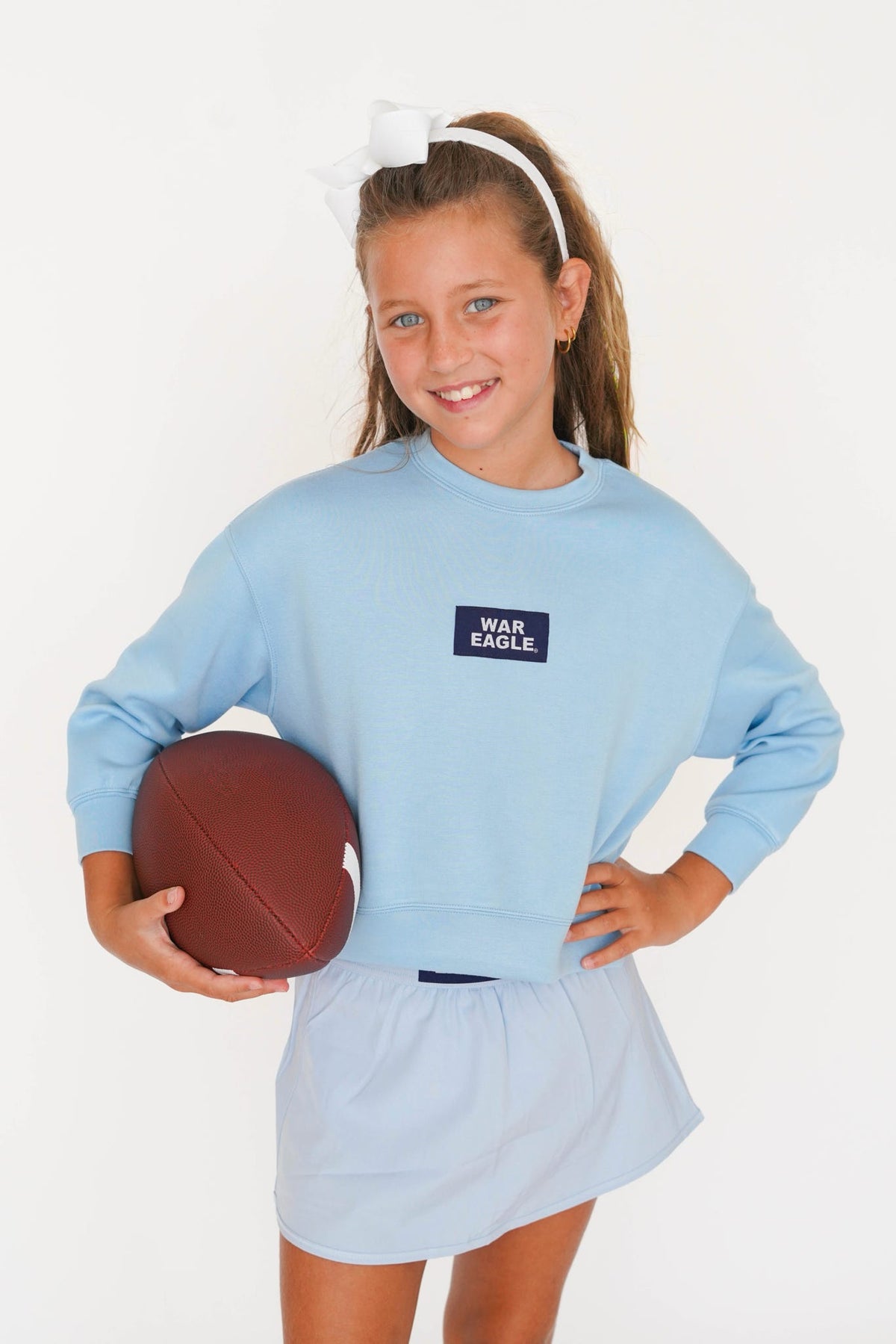 A young girl with a white hair bow and headband models a light blue sweatshirt and matching light blue skirt. She holds a brown football against a clean white studio background.