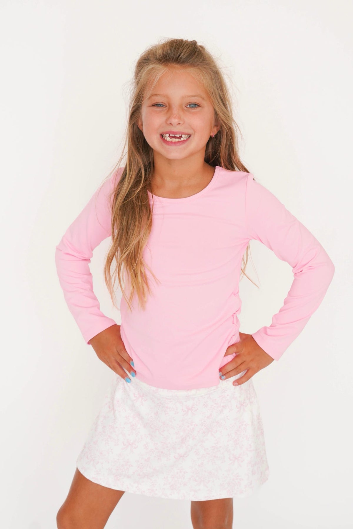 A young girl with long blonde hair smiling, wearing a light pink long-sleeve top and a white patterned skirt, posing with hands on hips against a plain white background.