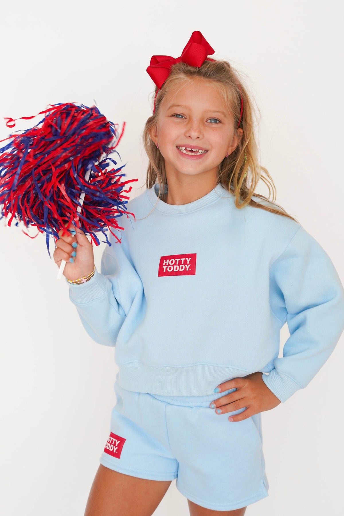 A young girl with blonde hair and a red bow is wearing a light blue sweatshirt and matching shorts set from Hotty Toddly, holding a red and blue pom-pom. The background is a clean white studio backdrop.