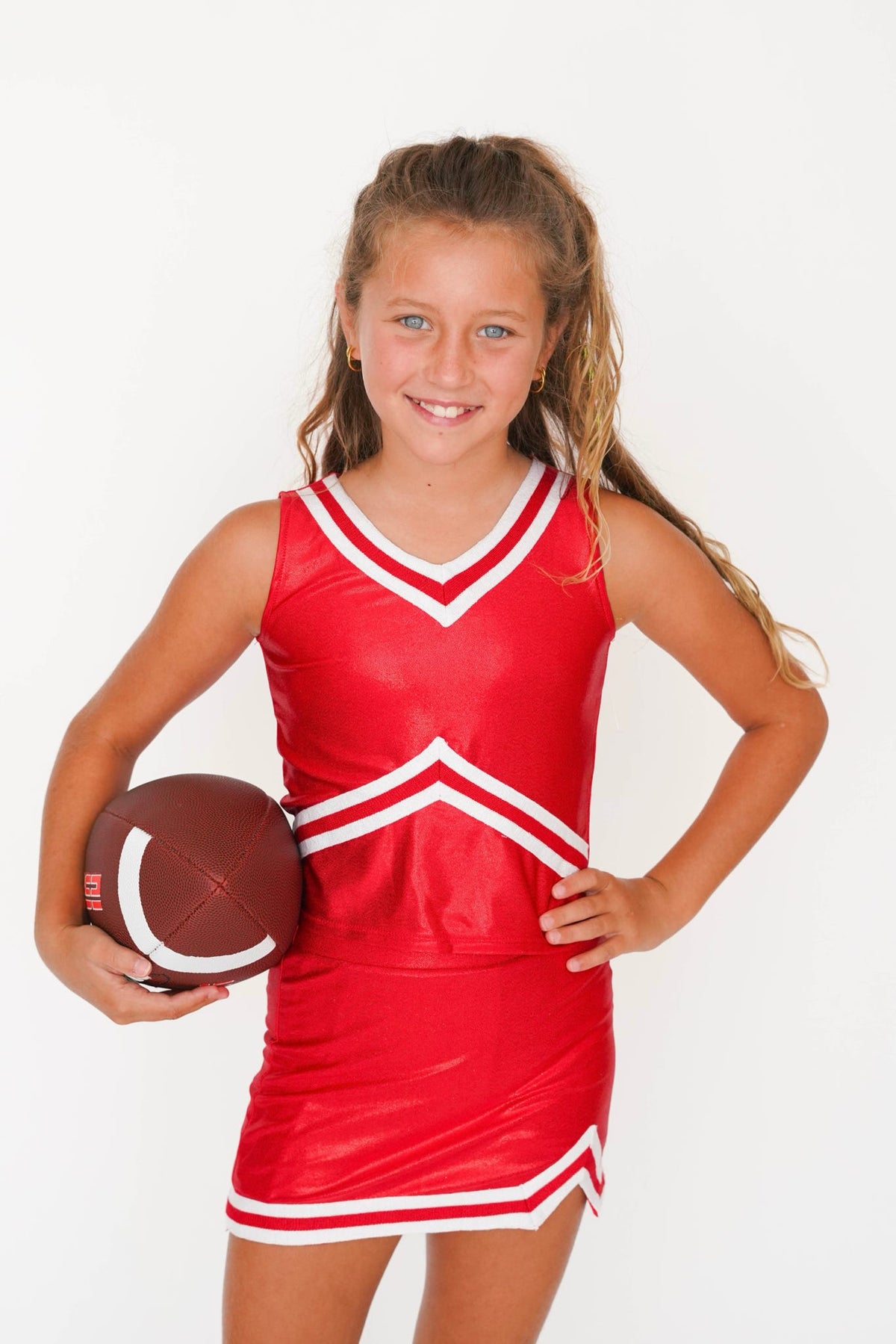 A young girl with blonde hair in a ponytail is wearing a shiny red cheerleader-style mini dress with white trim and holding a brown football against a plain white background.