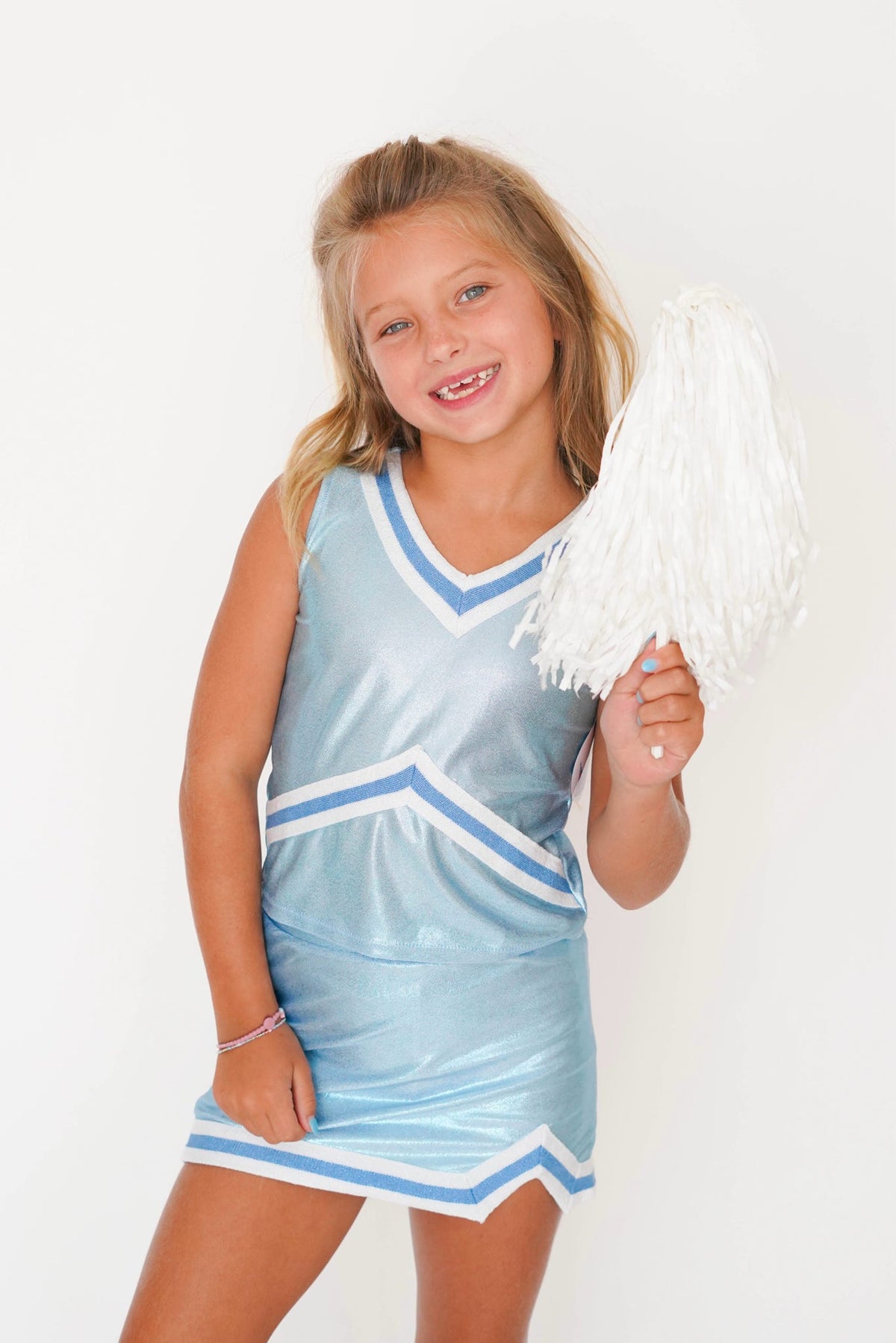 A young girl with blonde hair smiling and holding a white pom-pom, wearing a shiny light blue cheerleader dress with white and blue chevron stripes, set against a plain white background.