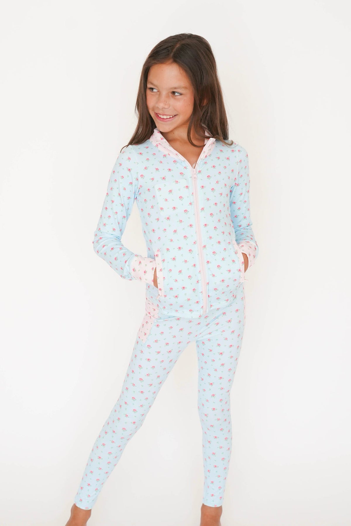 A young girl wearing a light blue long-sleeve zip-up pajama set with a pink and white pattern, standing against a plain white background.
